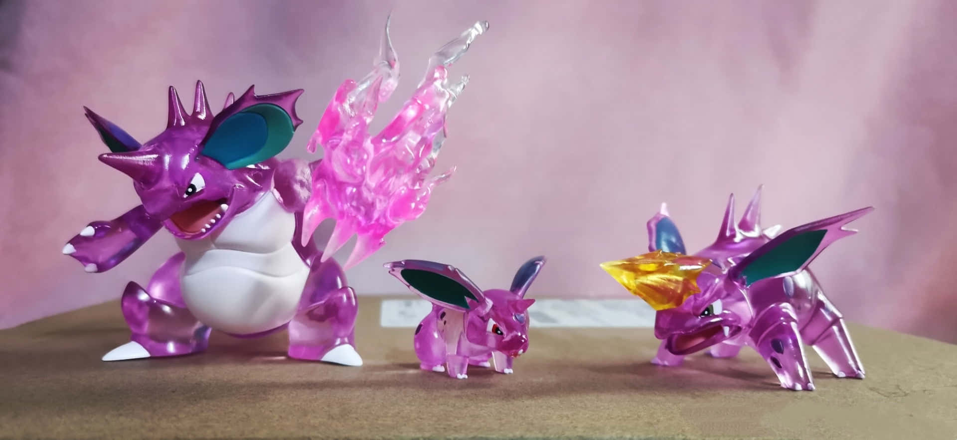 Nidoking 於其自然棲息地展現強大力量 壁紙 — 三個半透明紫色寶可夢公仔，帶有發光的粉紅色和黃色點綴，站在木質表面上，背景為柔和粉紅色。