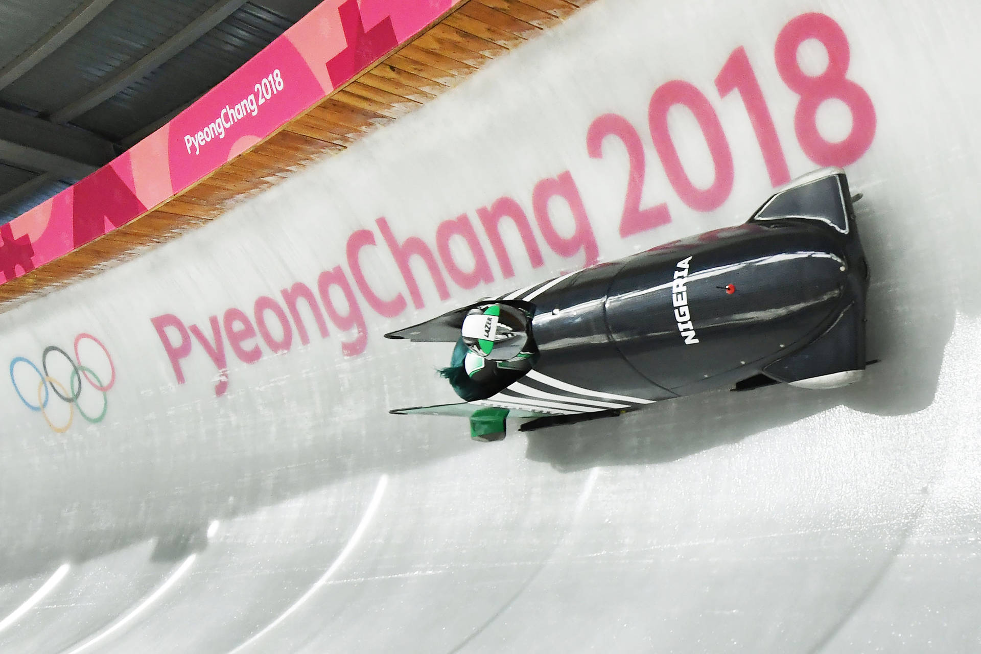 2018 平昌冬季奧運會 壁紙 — 尼日利亞雪車隊在平昌2018年冬季奧運會上沿著冰道飛馳，頭頂上有鮮豔的粉紅色品牌標誌。