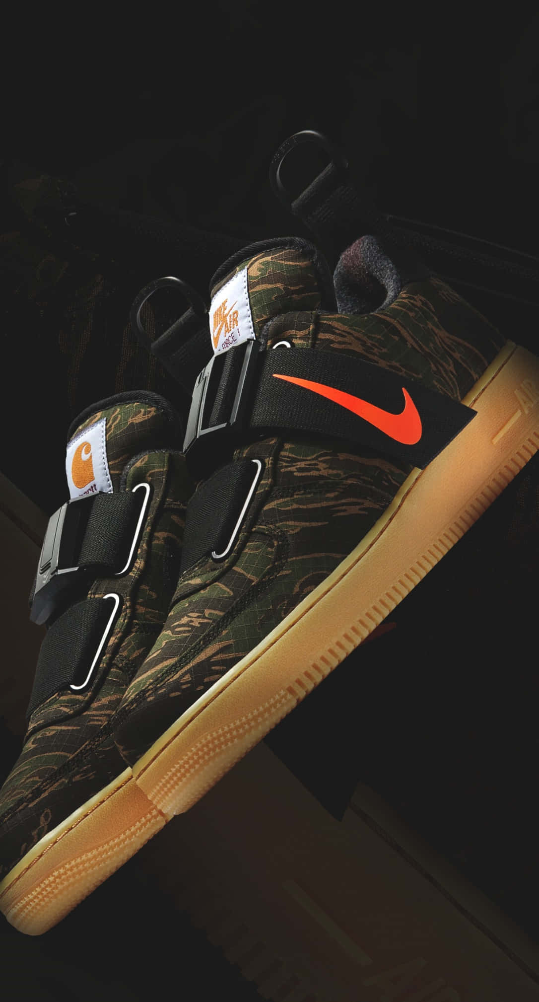 以Nike AF1展現街頭風格 壁紙 — 一對迷彩 Nike Carhartt 運動鞋，帶有醒目的橙色 swoosh 標誌和棕褐色鞋底，在深色背景下戲劇性地照亮。