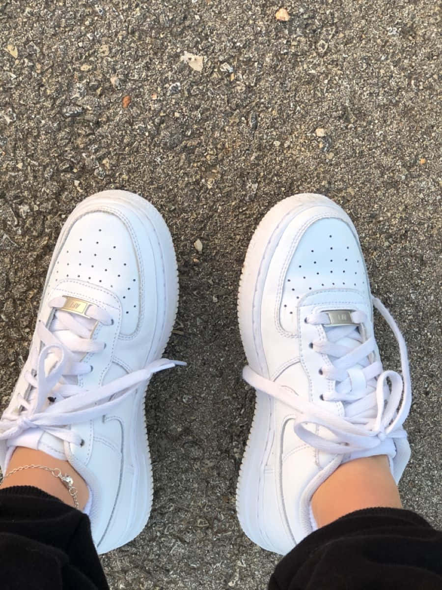穿著原版白色Nike Air Force 1的人圖 壁紙 — 帶有金色點綴的白色運動鞋從俯視角度放置在質感瀝青路面上。
