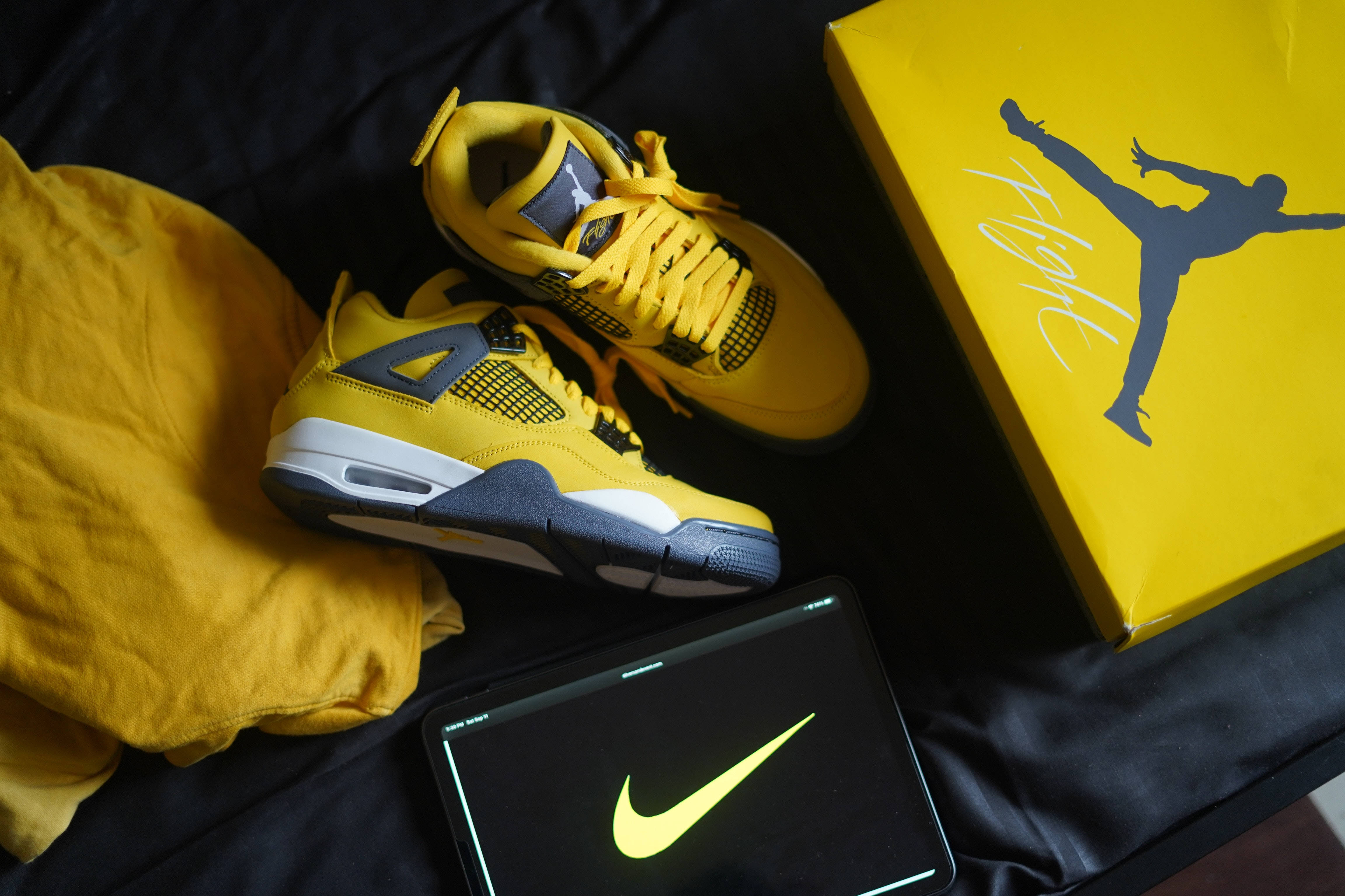 Nike Air Jordan 4 Retro 雷霆黃 壁紙 — 鮮黃色的Jordan 4運動鞋放在盒子旁邊，旁邊的平板電腦上顯示發光的Nike標誌，置於深色布料上。
