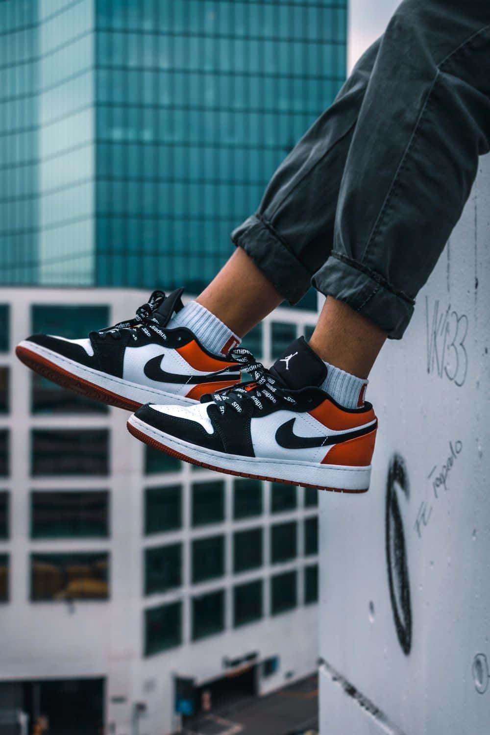 以 Nike Air Jordan 鞋款躍入全新風格 壁紙 — 一個人的腳穿著黑白橙色的運動鞋，隨意地掛在塗鴉牆上，背景是玻璃摩天大樓。