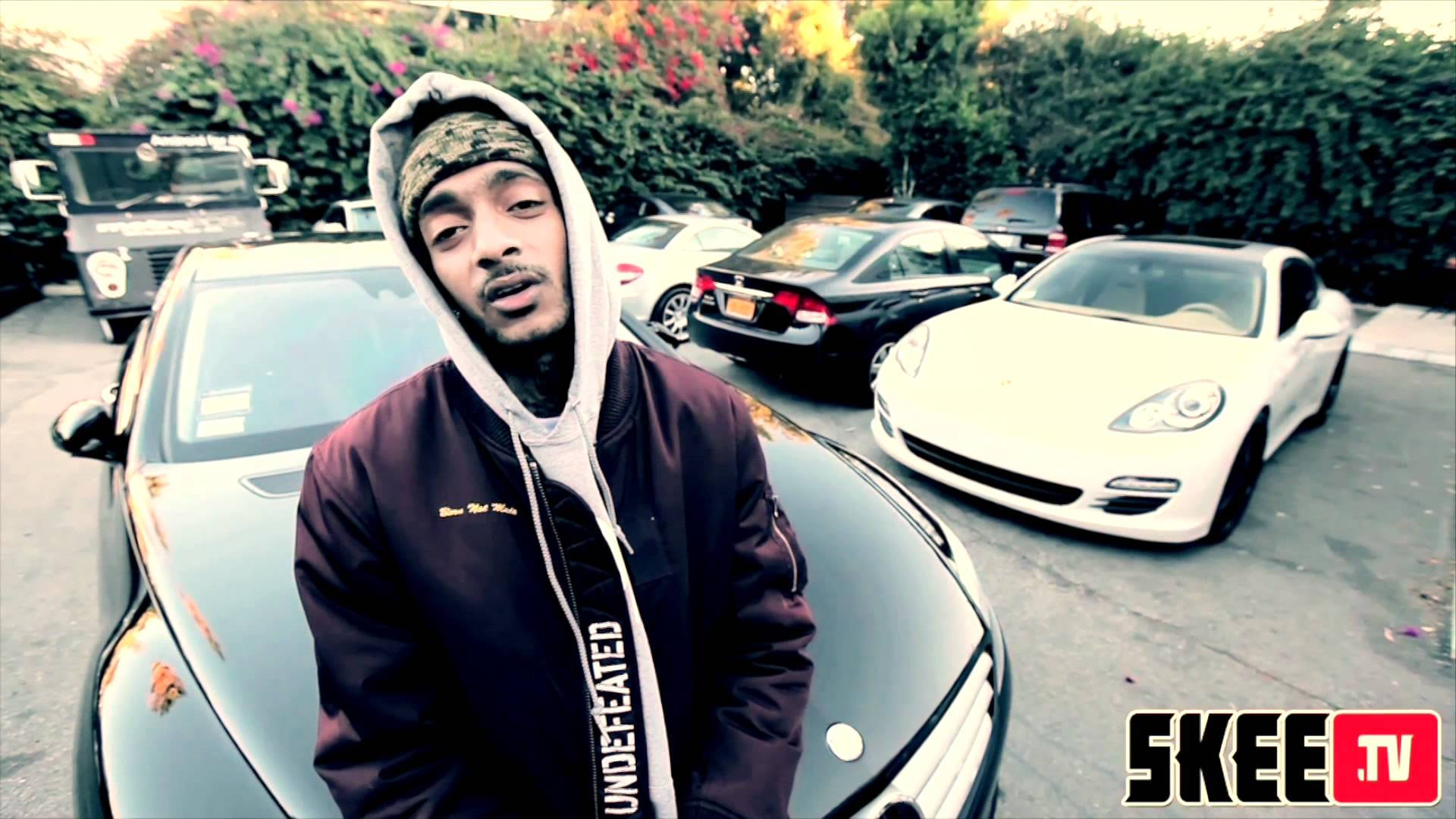 Nipsey Hussle —— 傳神姿態 壁紙 — 一名身穿連帽衫的男子自信地站在一輛黑色跑車旁邊，其他豪華車輛停放在陽光充足的停車場。