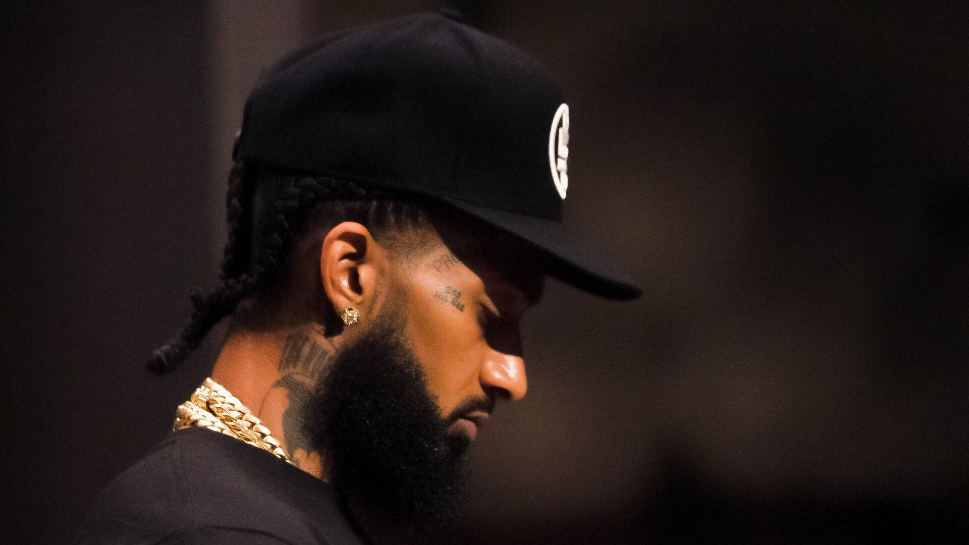 Nipsey Hussle 在放鬆時刻 壁紙 — 一個留鬍子、梳辮子的男人，在昏暗的環境中戴著黑色帽子和金鏈。