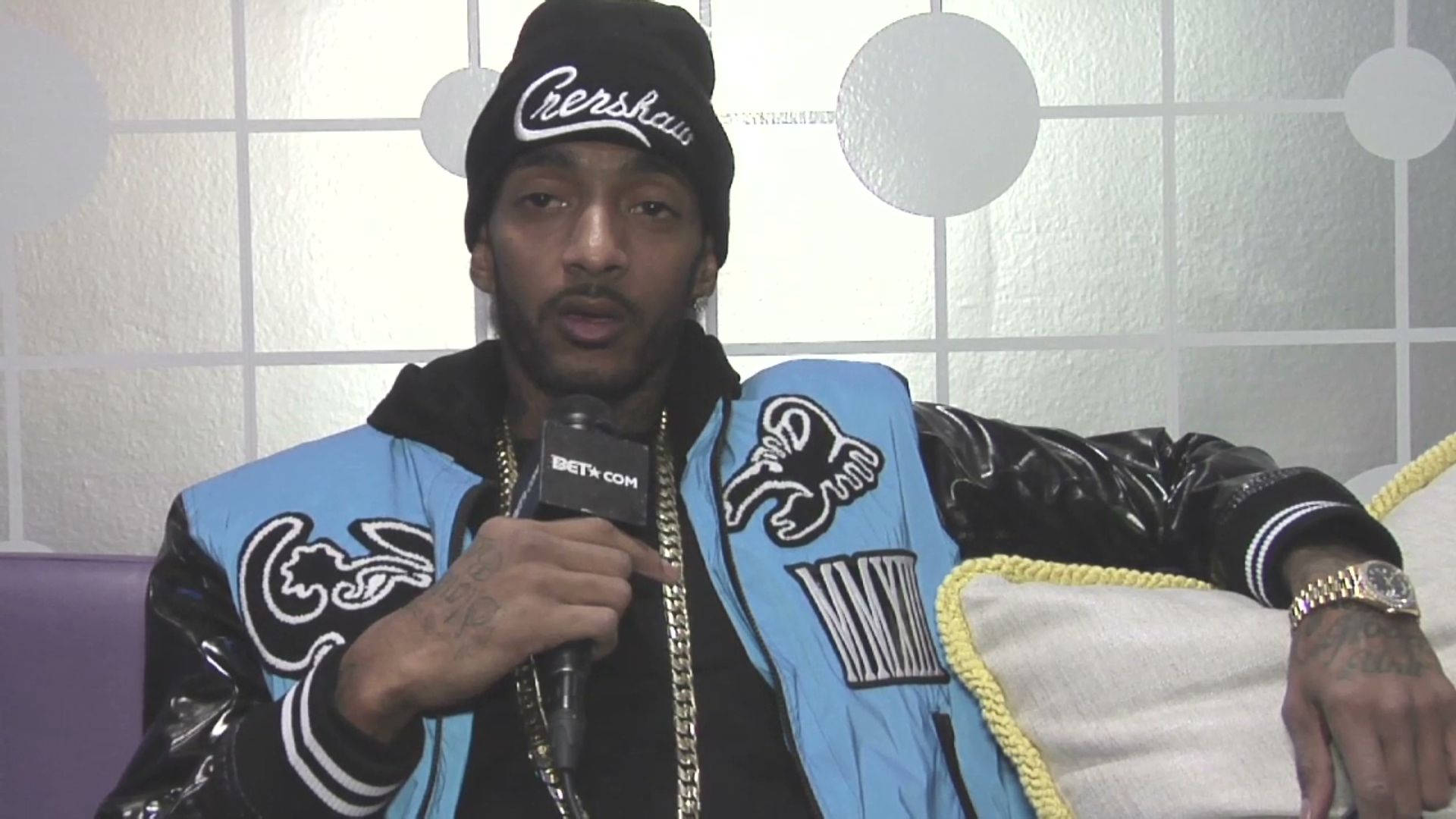 Nipsey Hussle 的深刻肖像 壁紙 — 一名身穿藍黑棒球外套和冷帽的男子，坐在瓷磚牆前的沙發上，手持麥克風。