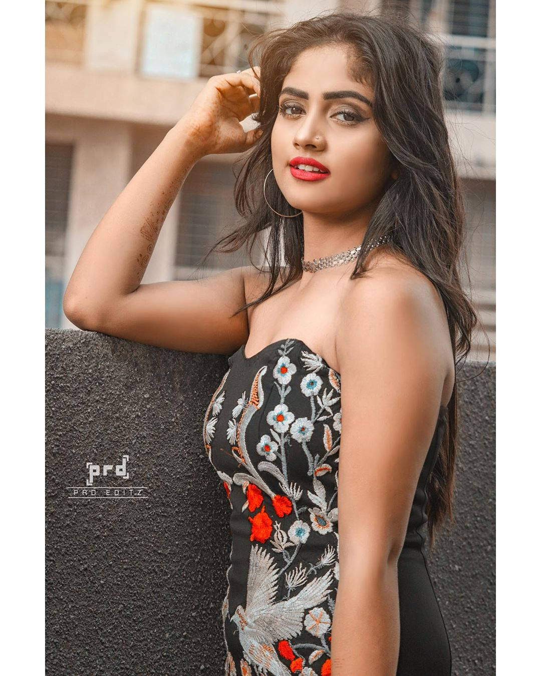 Nisha Guragain身穿花卉無袖上衣 壁紙 — 一位身穿黑色花卉刺繡連衣裙的女性，在柔和日光下，自信地以紋理牆壁和城市建築為背景擺姿勢。