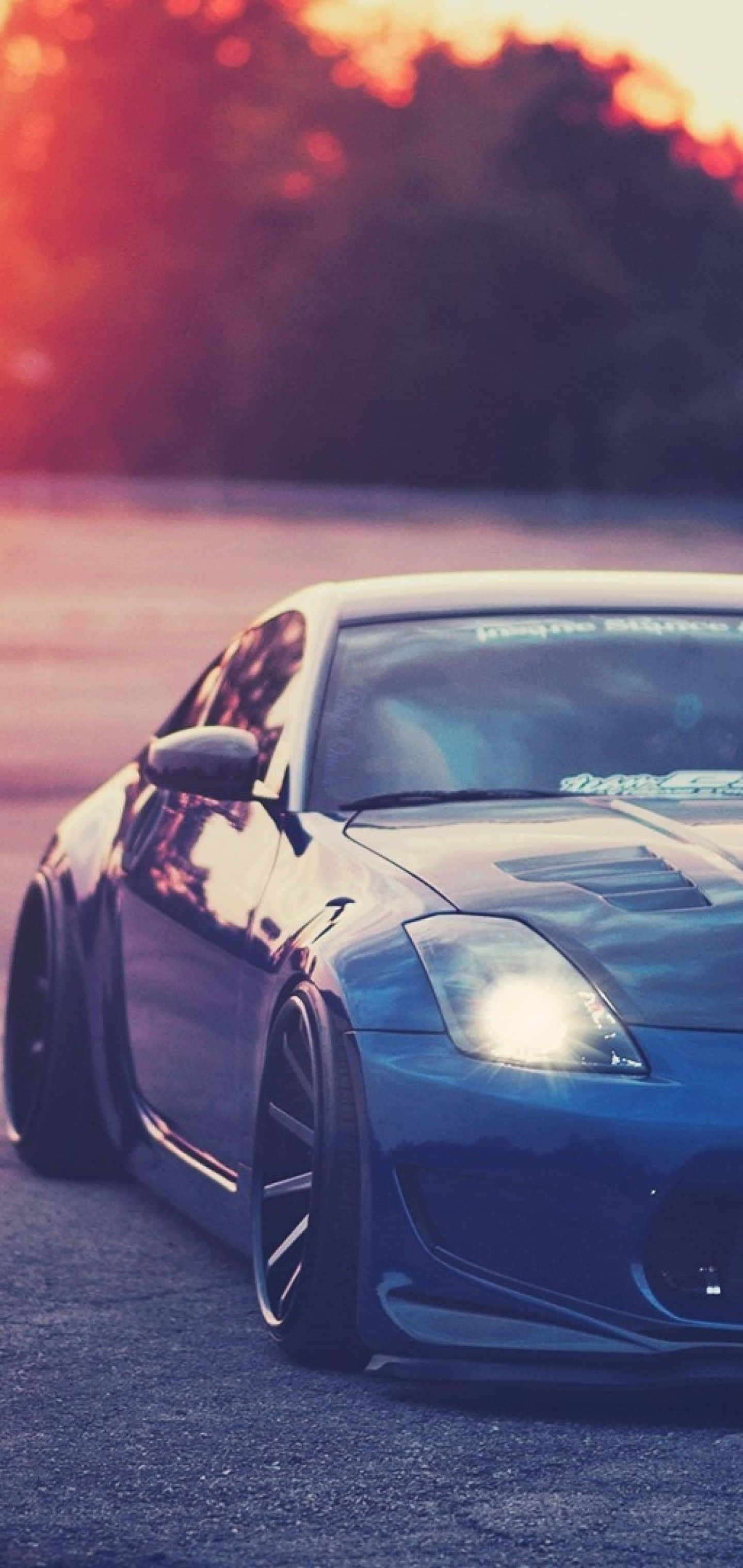 金屬藍 Nissan 350Z iPhone 美學照 壁紙 — 一輛時尚的藍色跑車在熾熱的日落天空下閃閃發光，背景是模糊的樹木。
