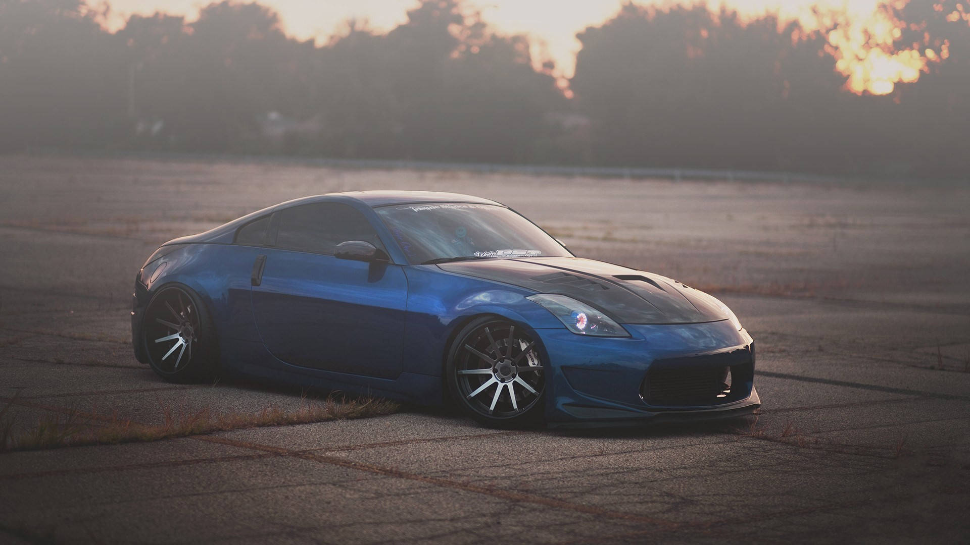 Nissan 350Z Z-Sport 運動跑車 壁紙 — 一輛時尚的藍色跑車帶有黑色點綴，低趴於裂開的瀝青地上，金色夕陽在樹後閃耀。