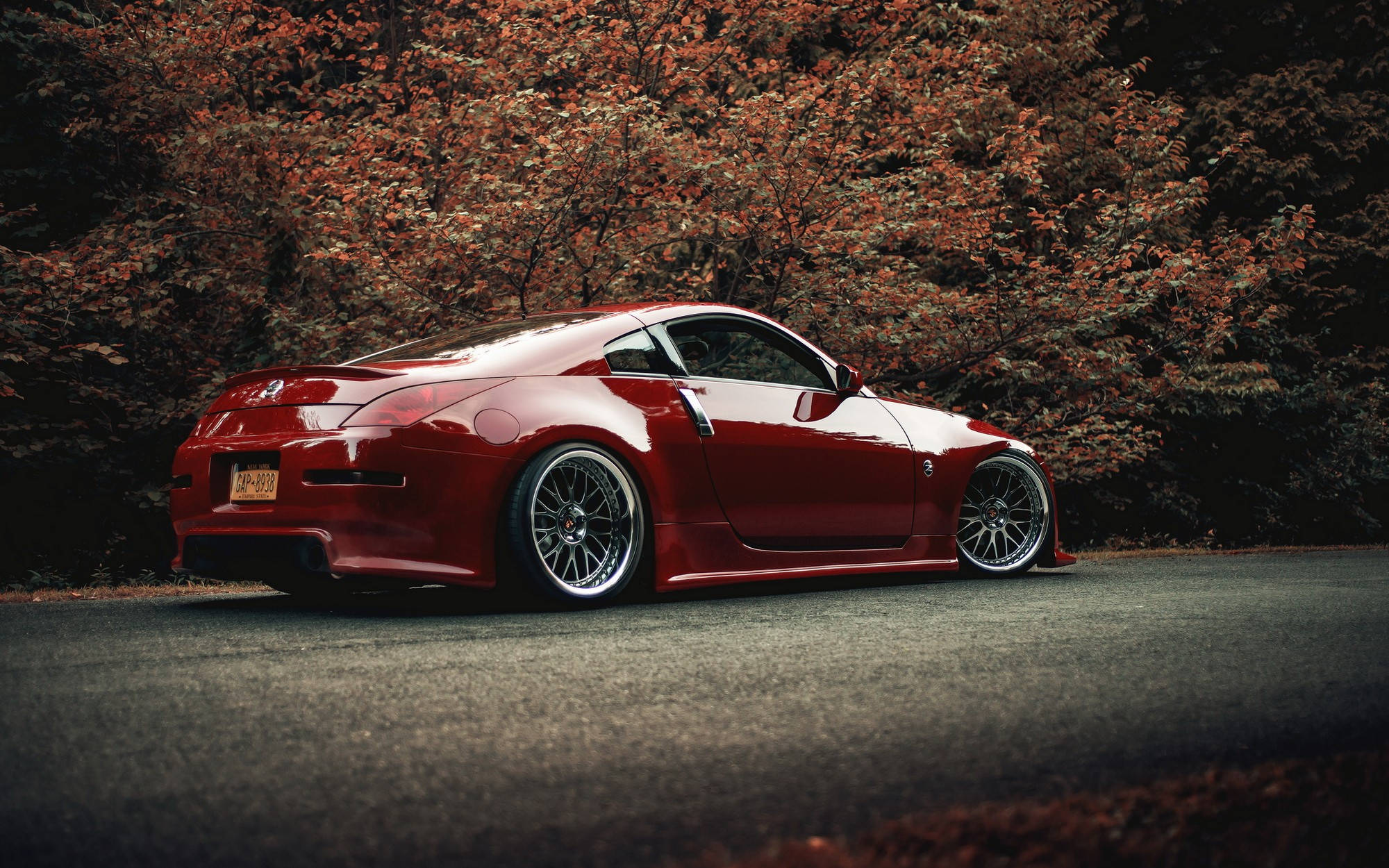 Nissan 350Z — 轉頭率最高的動力之選 壁紙 — 一輛光亮的紅色跑車配備定制輪圈，停在秋季樹葉覆蓋的瀝青路面上。