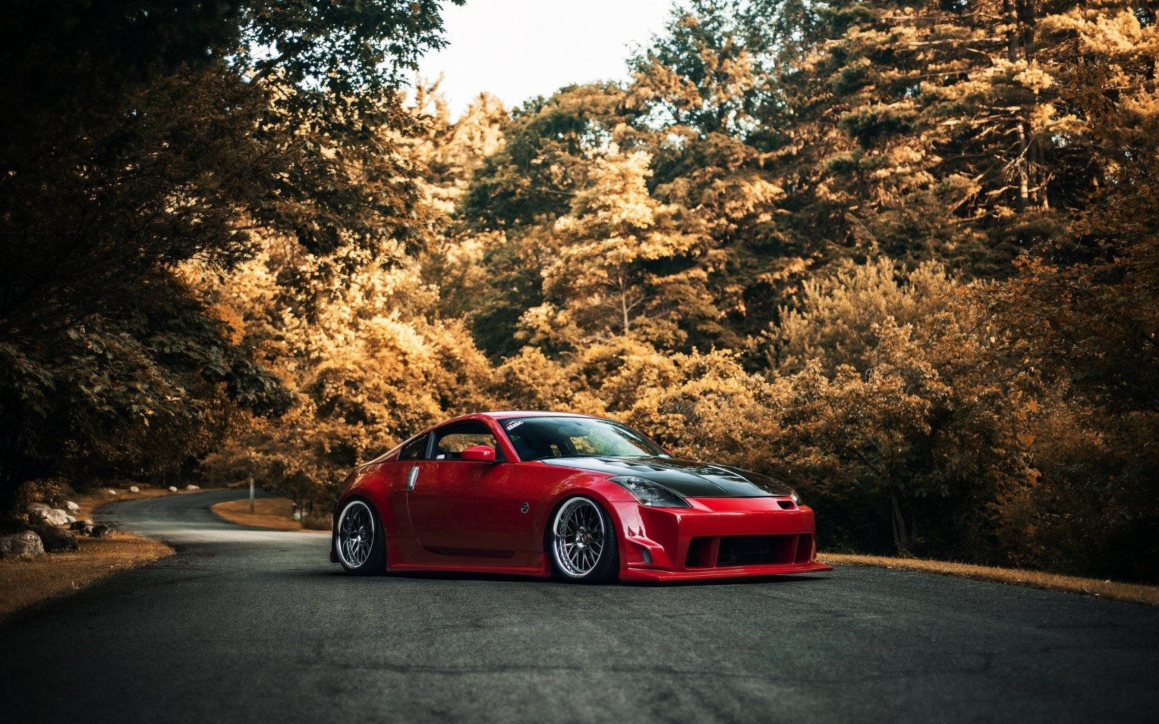 Nissan 350Z — 駛向無垠公路 壁紙 — 一輛帶有黑色飾邊的鮮紅色跑車停在蜿蜒的森林道路上，沐浴在金色的午後陽光中。