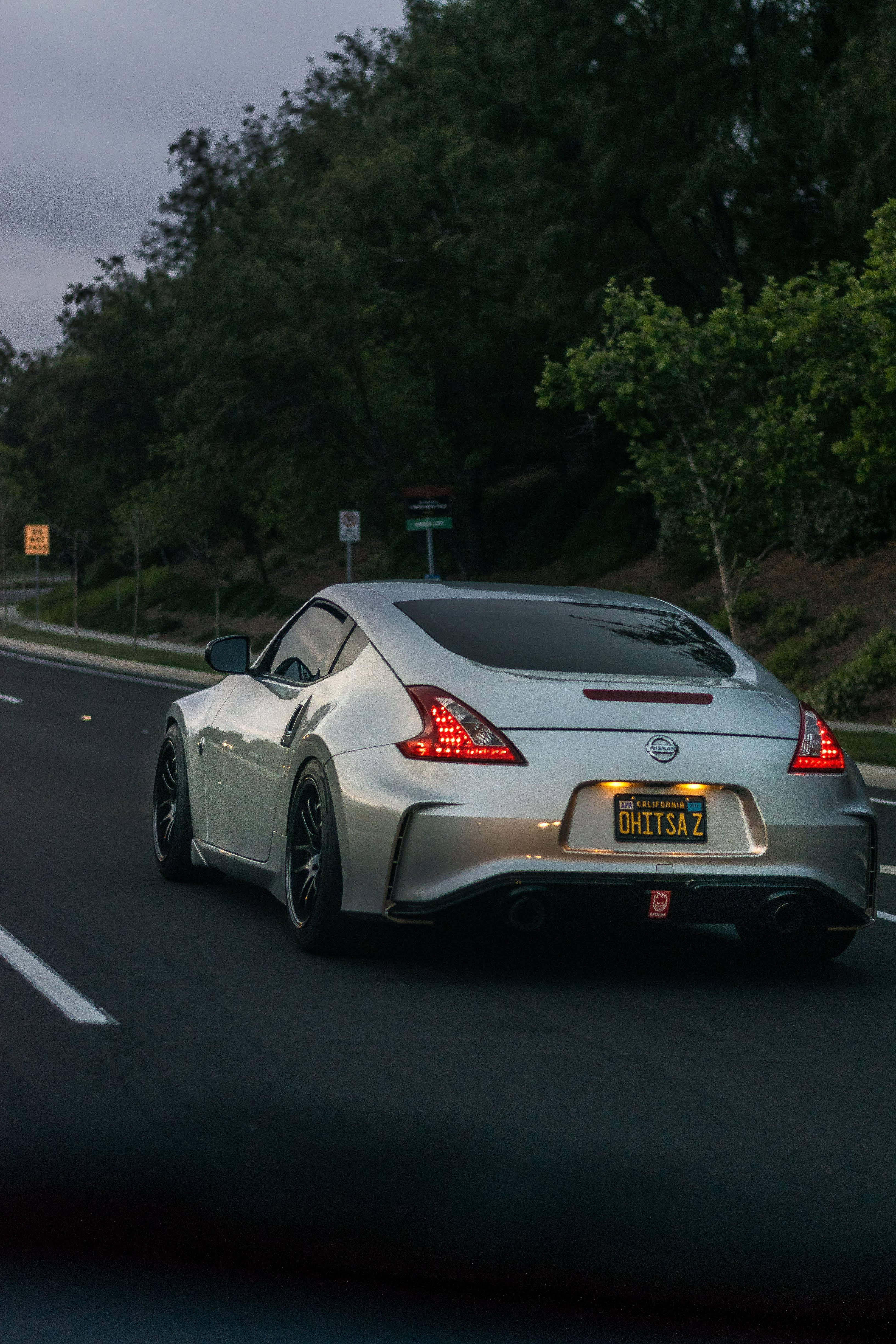 銀色 Nissan 370z 壁紙 — 一輛銀色跑車帶著發光的紅色尾燈，在黃昏時分行駛在綠樹成蔭的道路上。