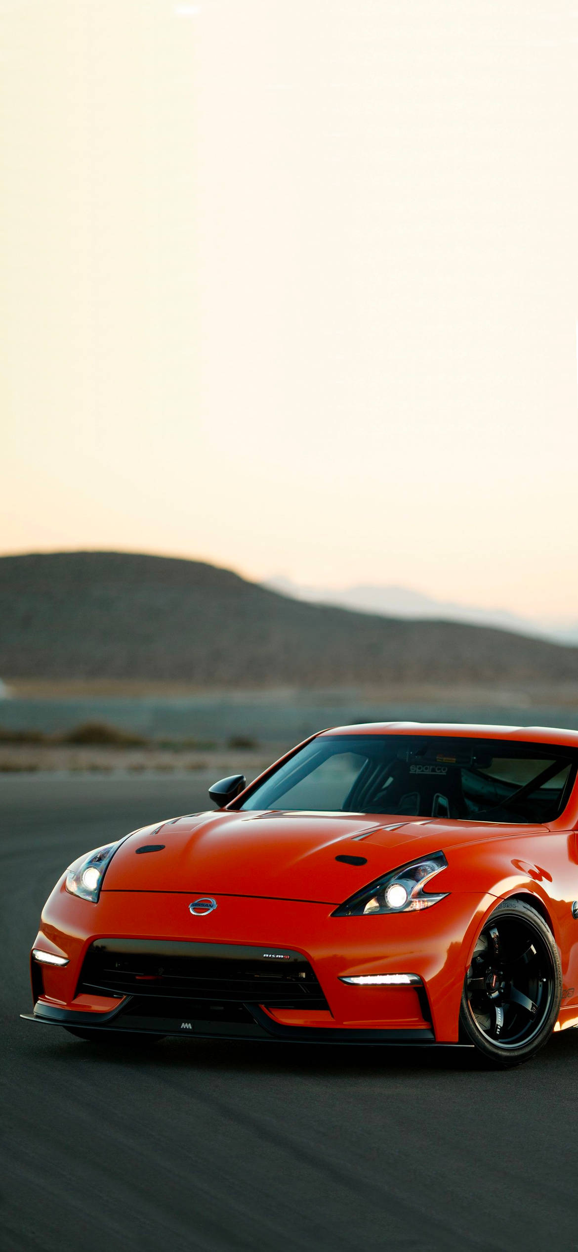 紅色Nissan 370Z與山丘 壁紙 — 一輛充滿活力的橙色跑車帶有黑色飾邊，在日落時分沿著沙漠公路疾馳，背景是遠山。