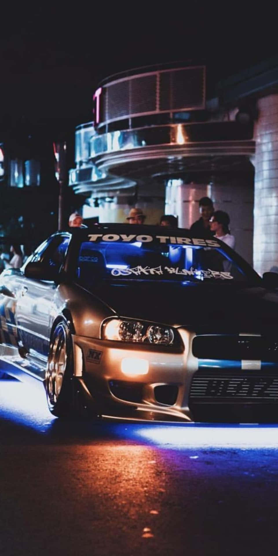 黑色改裝車帶有藍色底盤燈，夜間停車，有人觀看。