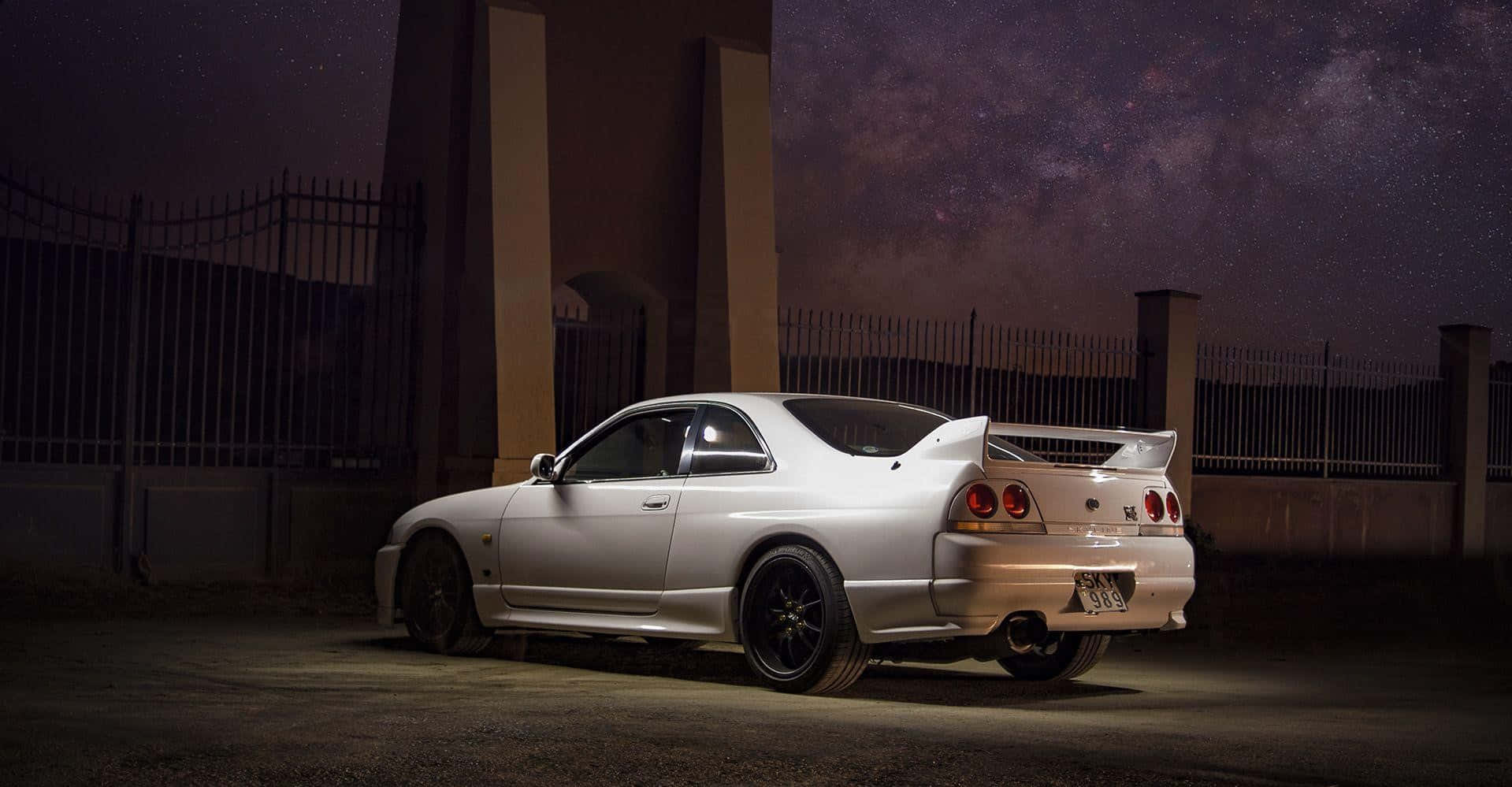 Nissan R33 GTR — 力量與掌控的化身 壁紙 — 一輛銀色跑車在聚光燈下閃閃發光，背景是繁星點點的夜空，籬笆和建築物上方可見銀河。