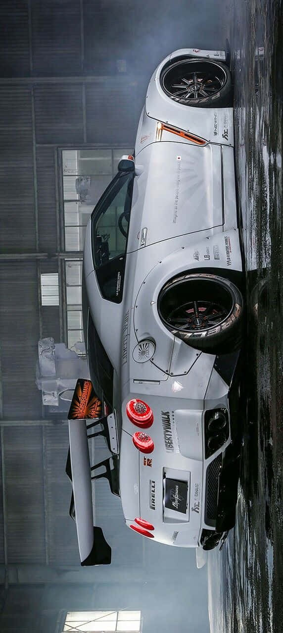 經典Nissan R35 GT-R 壁紙 — 一輛帶有紅色點綴和贊助商標誌的白色賽車停放在昏暗的工業車庫中，地面濕滑，蒸汽升騰。