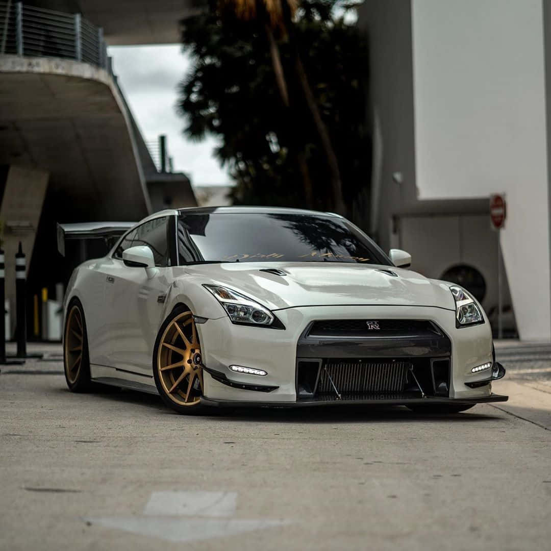 Nissan R35 GT-R 的流線美學 壁紙 — 一架時尚的白色日產GT-R配金色輪圈，停泊在現代混凝土天橋下，背景有棕櫚樹。