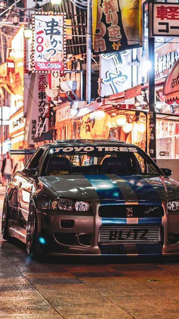 Nissan R35 GT-R 街頭圖 壁紙 — 一輛經過改裝的銀色跑車帶有藍色條紋，夜晚停在潮濕的霓虹燈日本街道上。