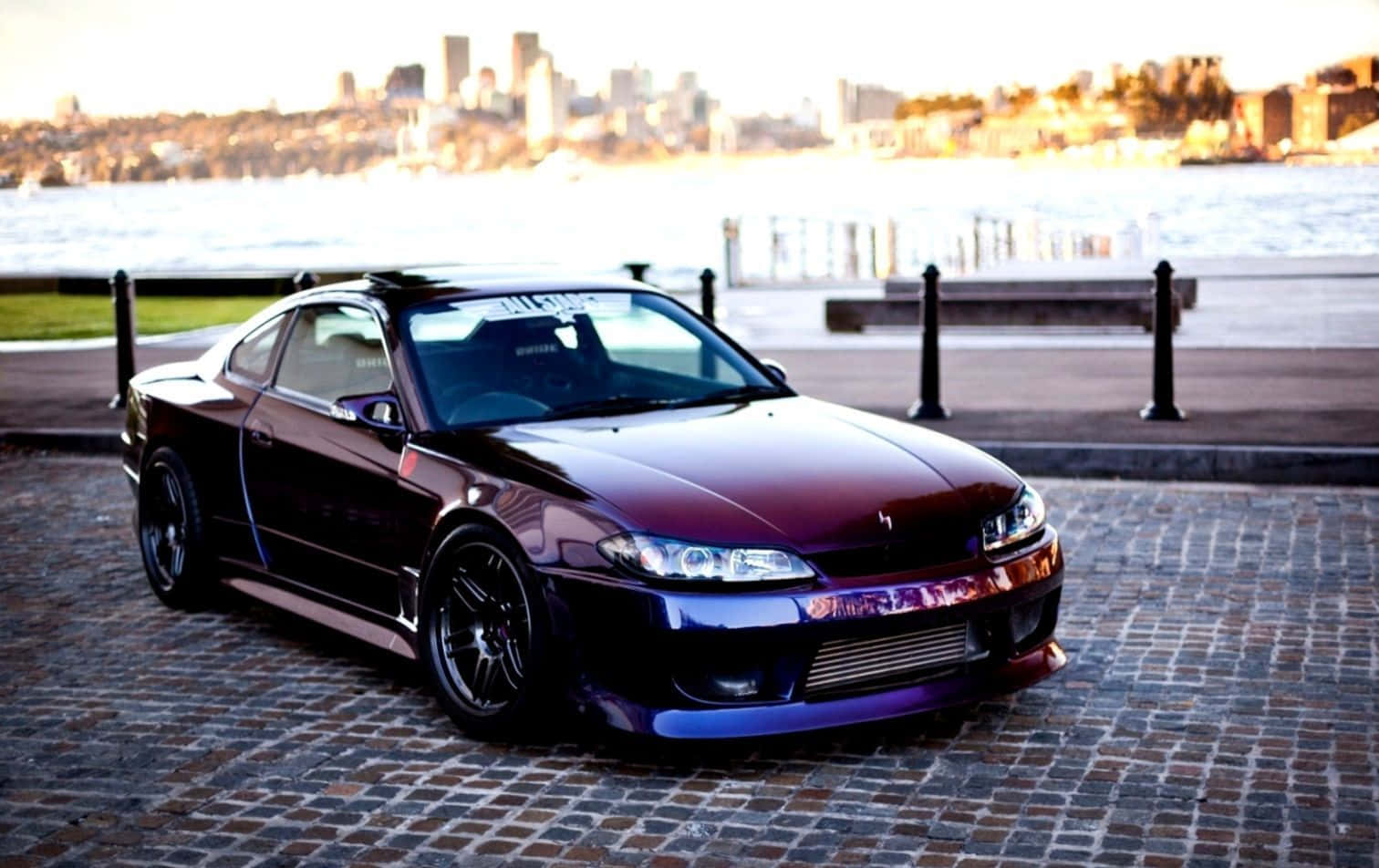 Nissan Silvia S15 活力紅款 壁紙 — 一輛光澤栗色跑車配備定制輪轂，停泊在鵝卵石海濱，正值黃金時段，背景是城市天際線。