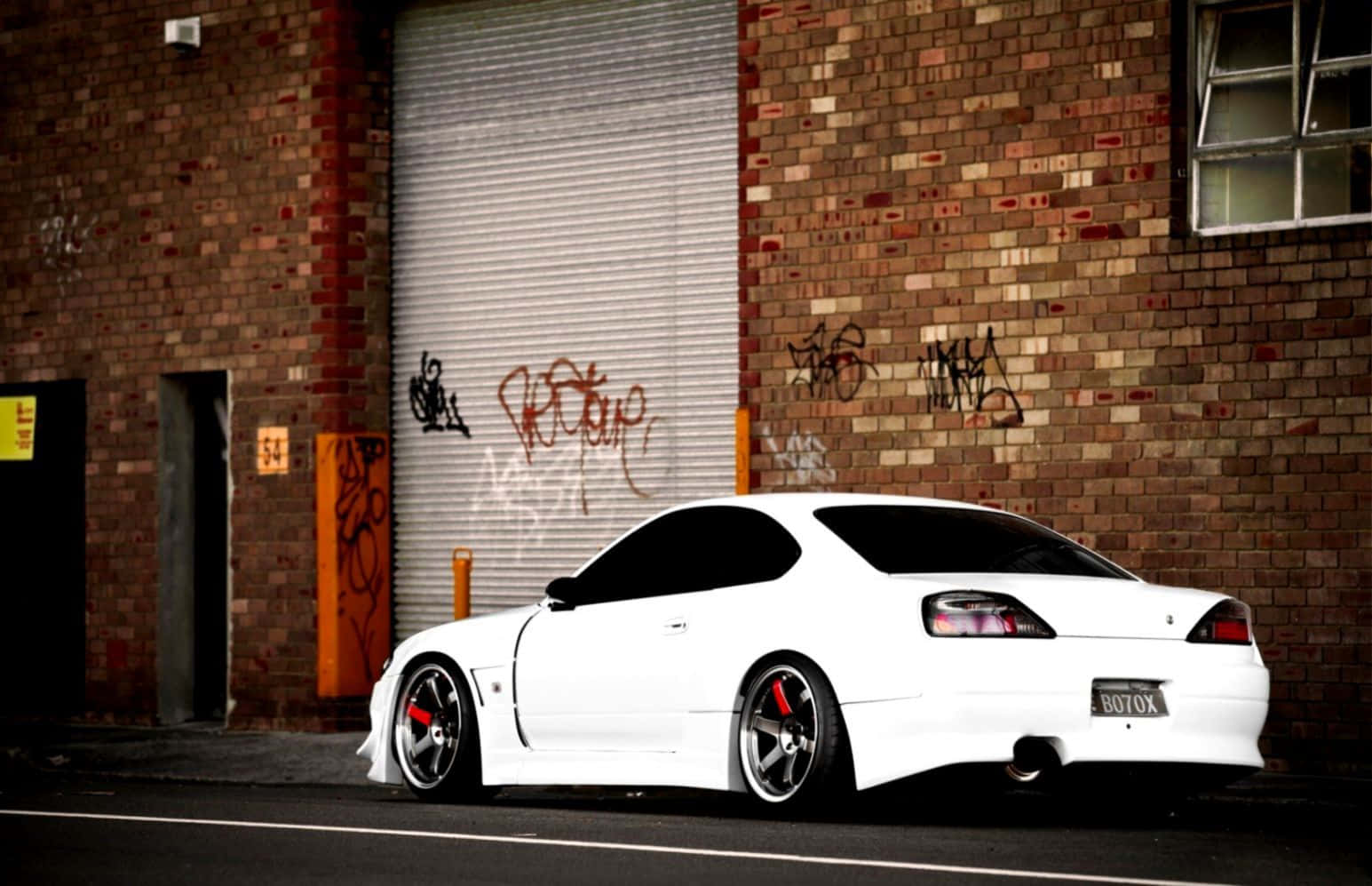 Nissan Silvia S15 與塗鴉磚牆 壁紙 — 一輛帶有紅色制動卡鉗的純白色跑車停在一個佈滿塗鴉的磚砌建築和金屬捲簾門前。