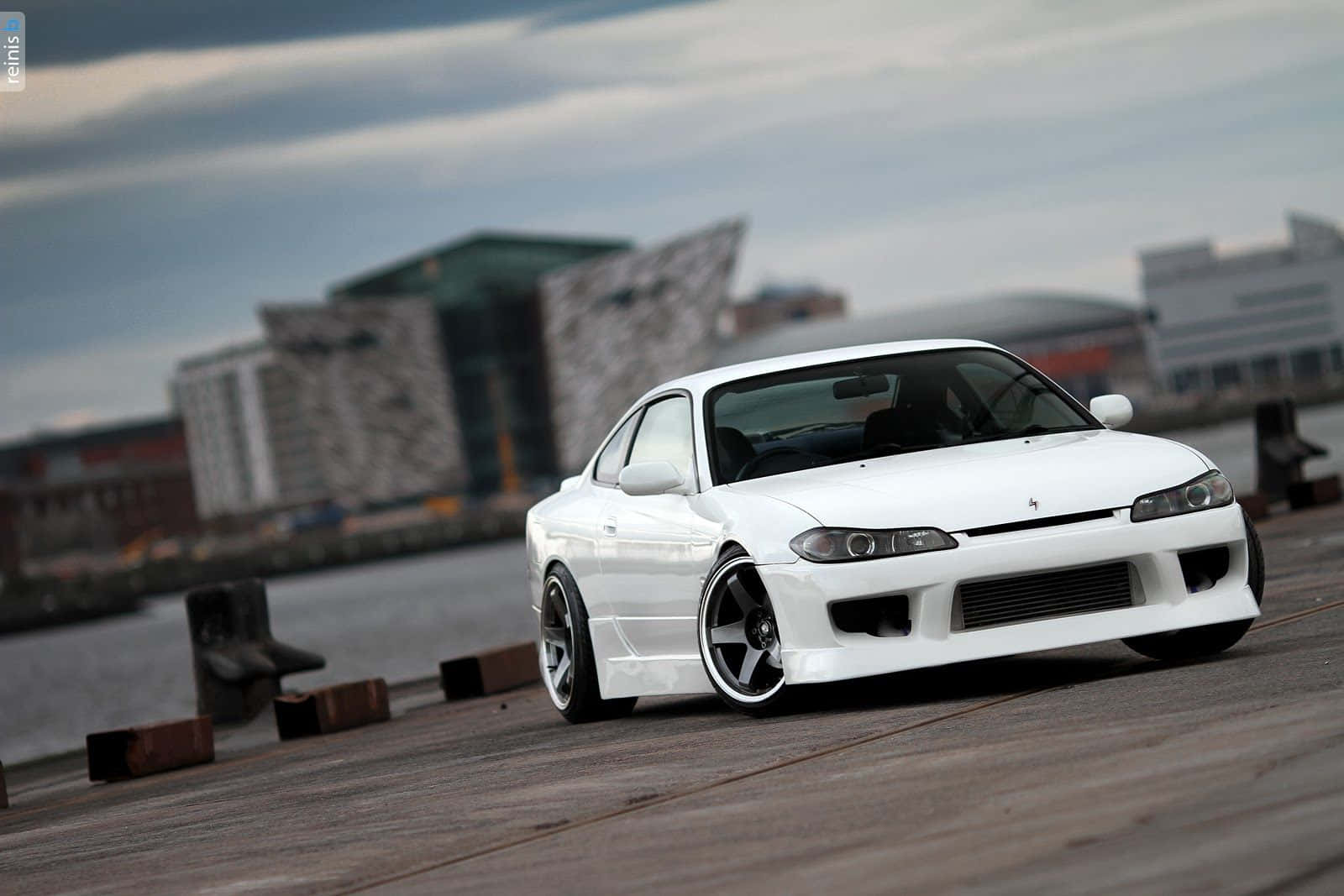 Nissan Silvia S15 工業園區風景 壁紙 — 一架時尚的白色跑車配備定制輪圈，停泊在木製碼頭旁，背景是現代建築，天空多雲。
