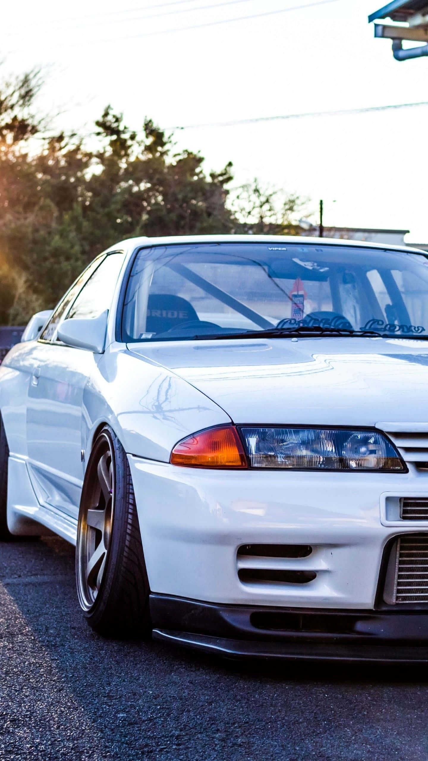 標題：震撼的動力——經典Nissan Skyline 壁紙 — 一輛低矮的白色跑車，配備定制輪轂和賽車貼花，在瀝青路上的金色夕陽下閃閃發光。