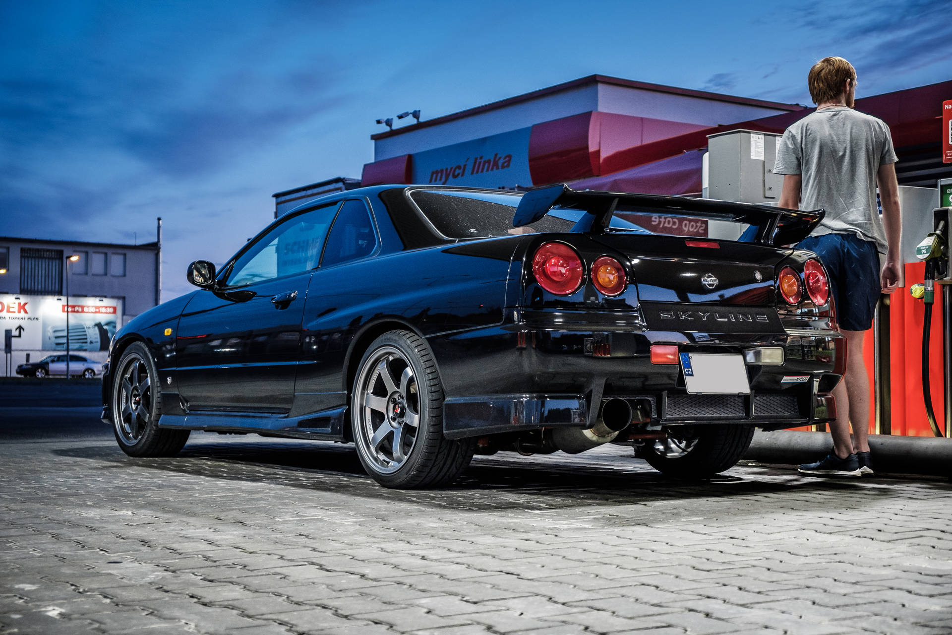 Nissan Skyline GTR R34 跑車 壁紙 — 一架亮黑色日產Skyline R34停泊在加油站，一名男子在暮色下為其加油。