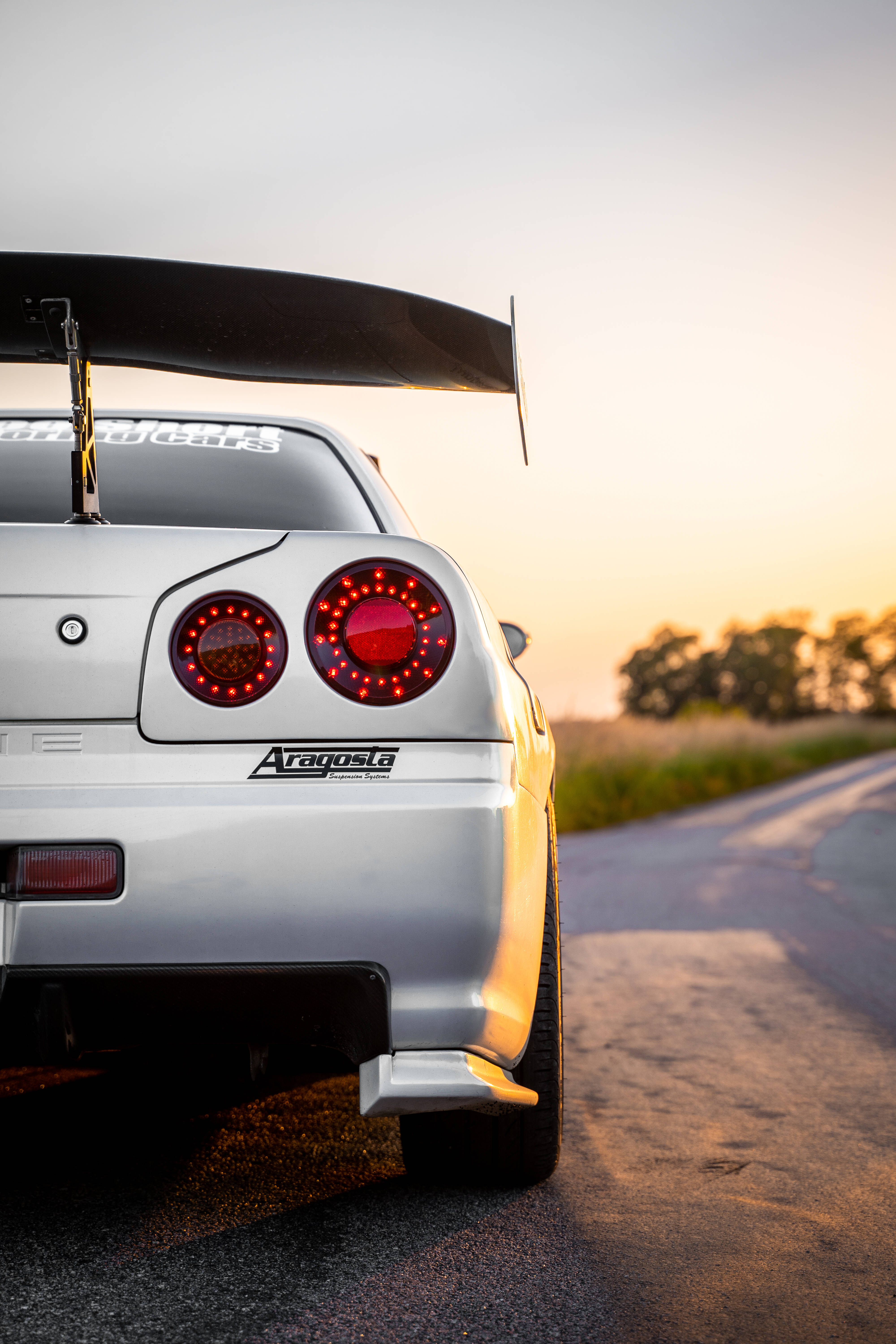 Nissan Skyline GTR R34 半身後視 壁紙 — 一輛銀色日產GT-R，帶有發光的紅色尾燈和大型後擾流板，停在日落時分的鄉村道路上。