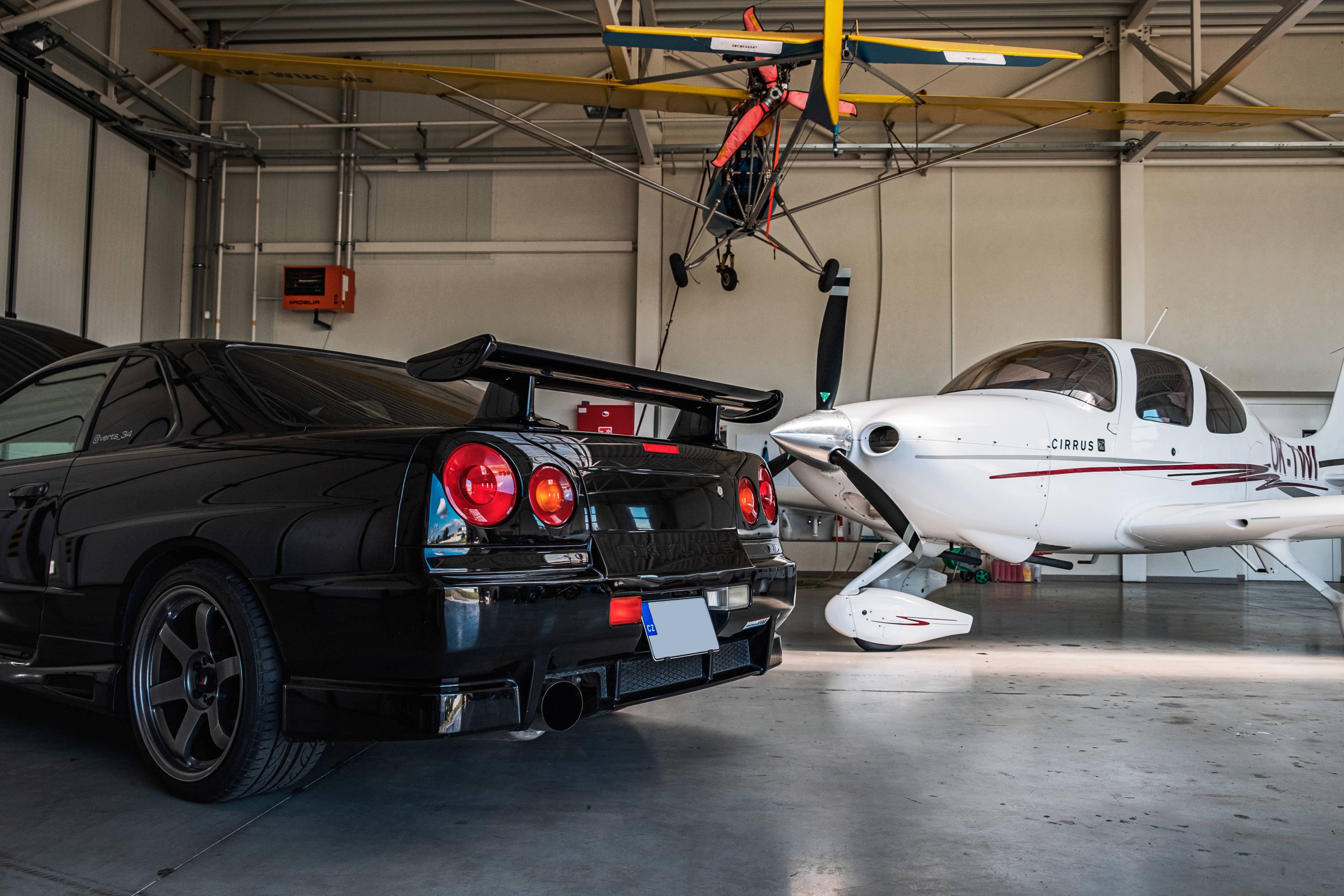 Nissan Skyline GTR R34 機庫 壁紙 — 一架時尚黑色跑車和一架白色單引擎飛機共用一個寬敞的機庫，上方懸掛著一架黃色旋翼機。