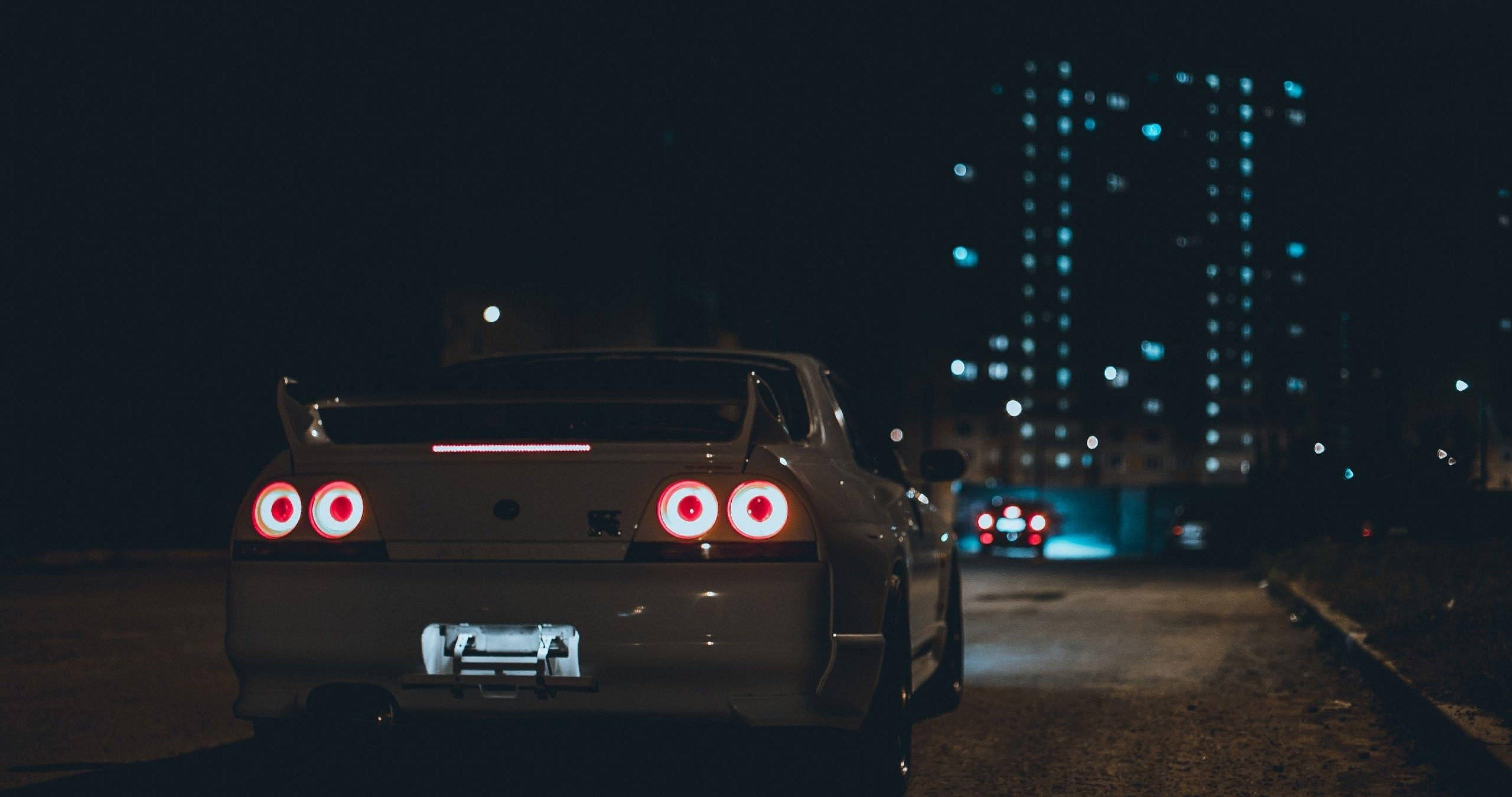Nissan Skyline GTR R33 後視圖 壁紙 — 一輛銀色跑車在夜間停泊在黑暗的城市街道上，紅色尾燈閃爍，背景是燈火通明的摩天大樓。