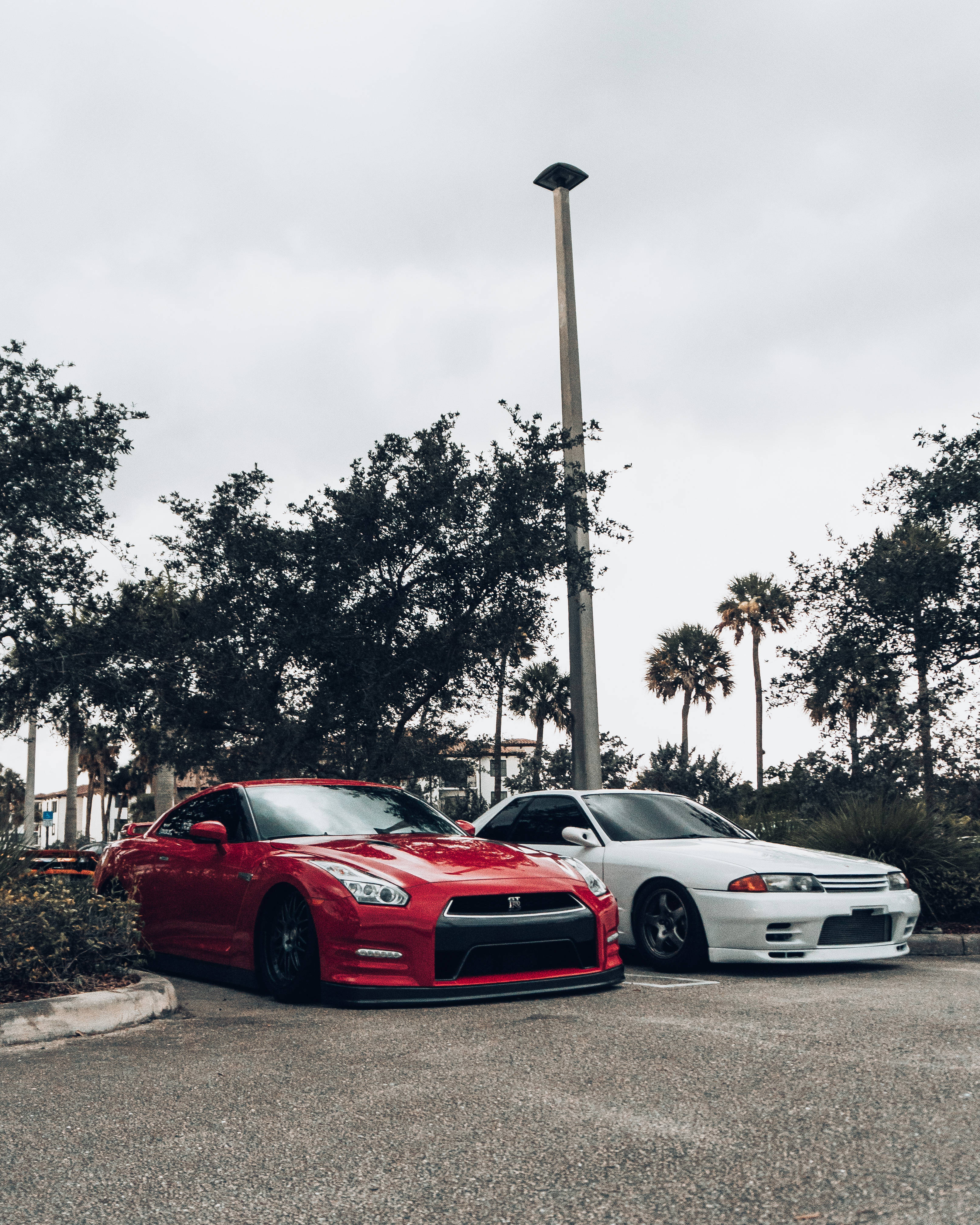 改裝版紅白 Nissan Skyline GTR R34 壁紙 — 兩輛時尚跑車，一輛鮮紅色的日產GT-R和一輛純白色的日產Skyline，並排停放在陰天的停車場，周圍環繞著棕櫚樹和茂密的綠色植物。