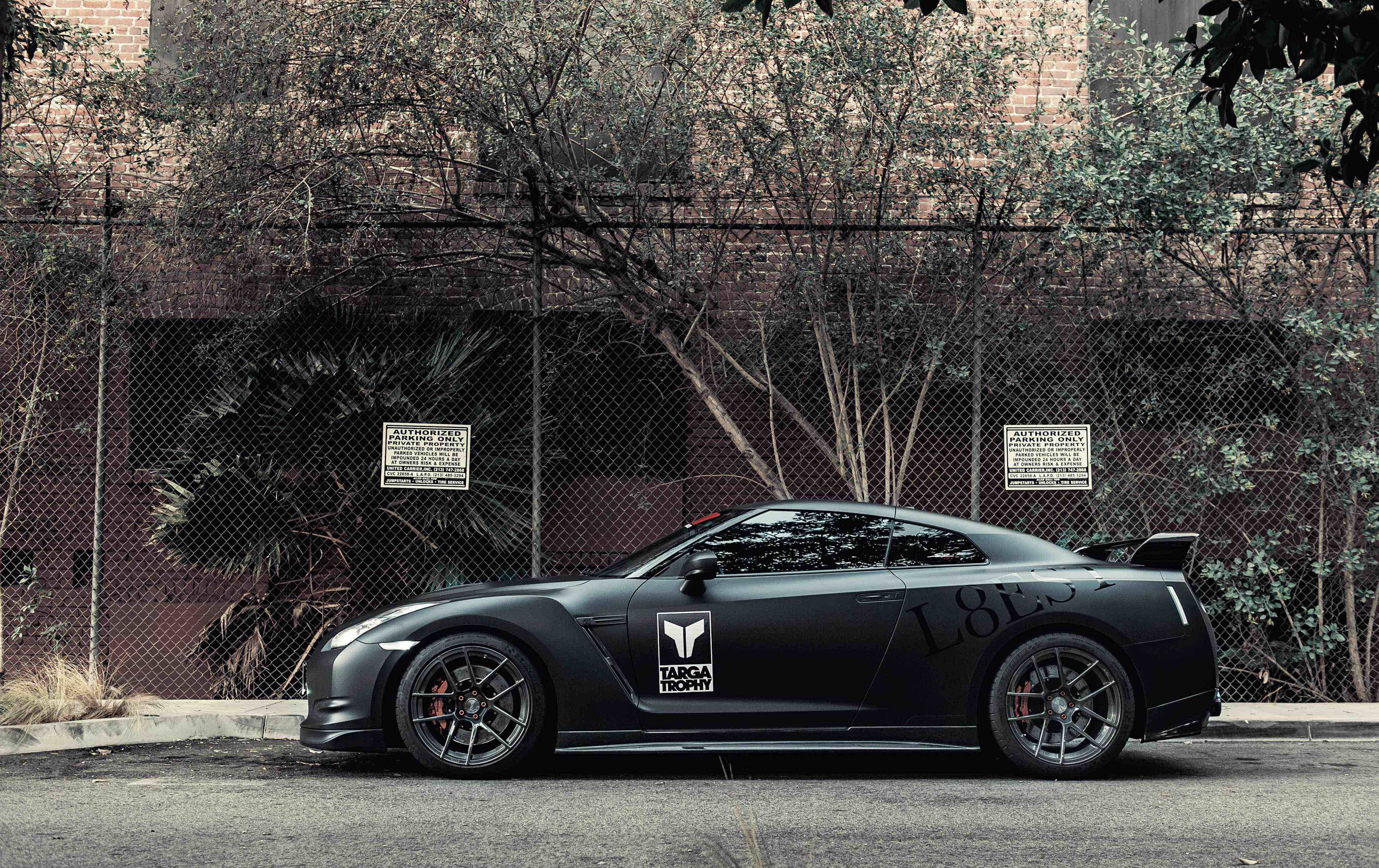 標題：強勁的Nissan Skyline GTR R35停在金屬網圍欄後 壁紙 — 一輛啞光黑色的日產GT-R跑車停泊在鐵絲網圍欄旁邊，背景有樹木和磚砌建築。