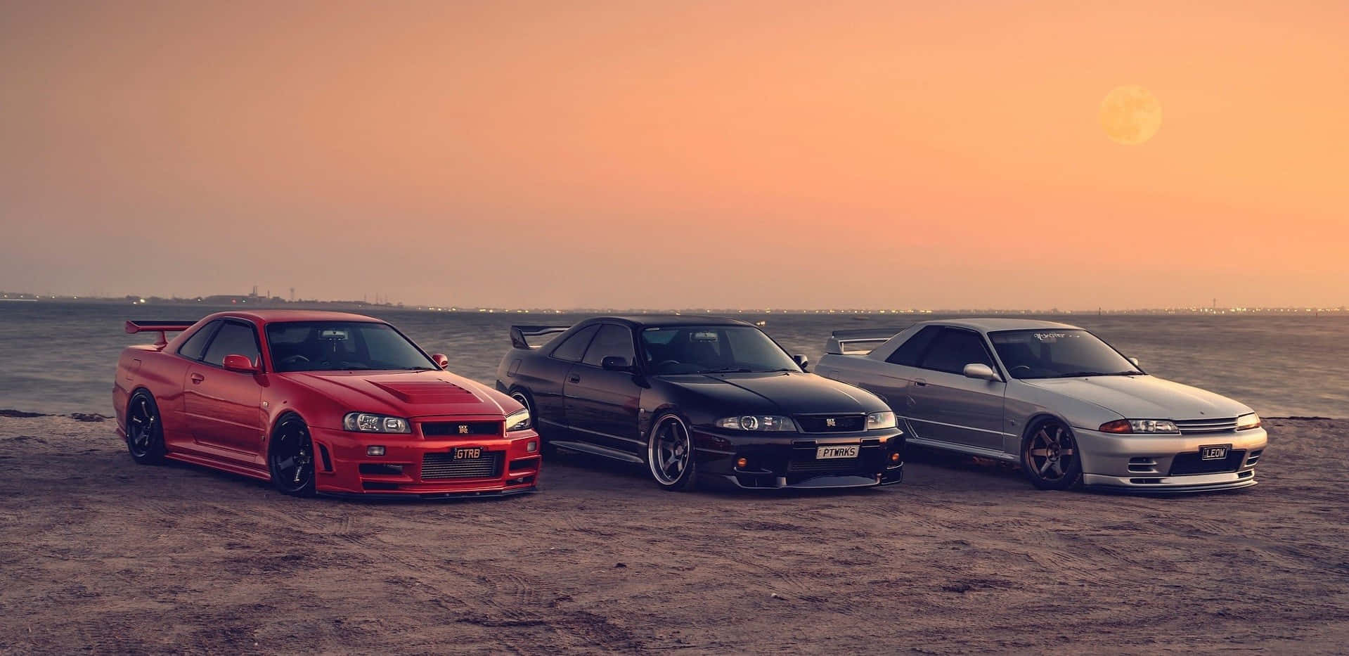 Nissan Skyline R33 GT-R 等待閃耀時刻 壁紙 — 三輛改裝日產Skyline GT-R停泊在日落時分的沙灘上，上方有一輪發光的月亮。