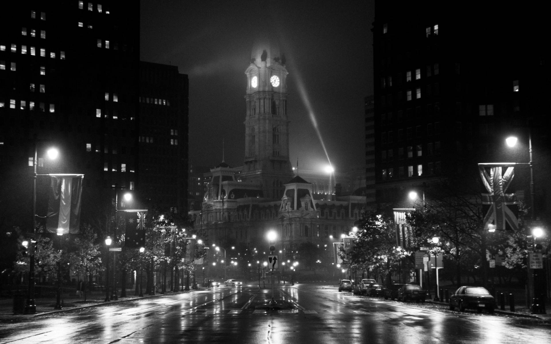 費城夜色 noir 風格 壁紙 — 黑白夜景城市景觀，特色係一個宏偉嘅鐘樓被聚光燈照亮，下方係一條濕漉漉、有倒影嘅街道，兩旁排列著建築物同街燈。