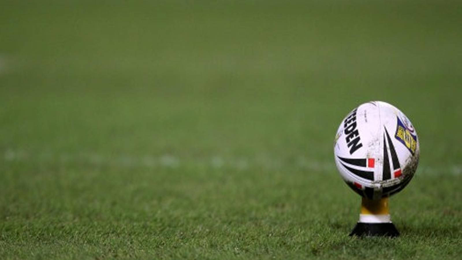 NRL 足球賽激烈進行中 壁紙 — 一個帶有黑色和黃色標記的白色欖球直立於一個充滿活力的綠色草地場地上的球座上。