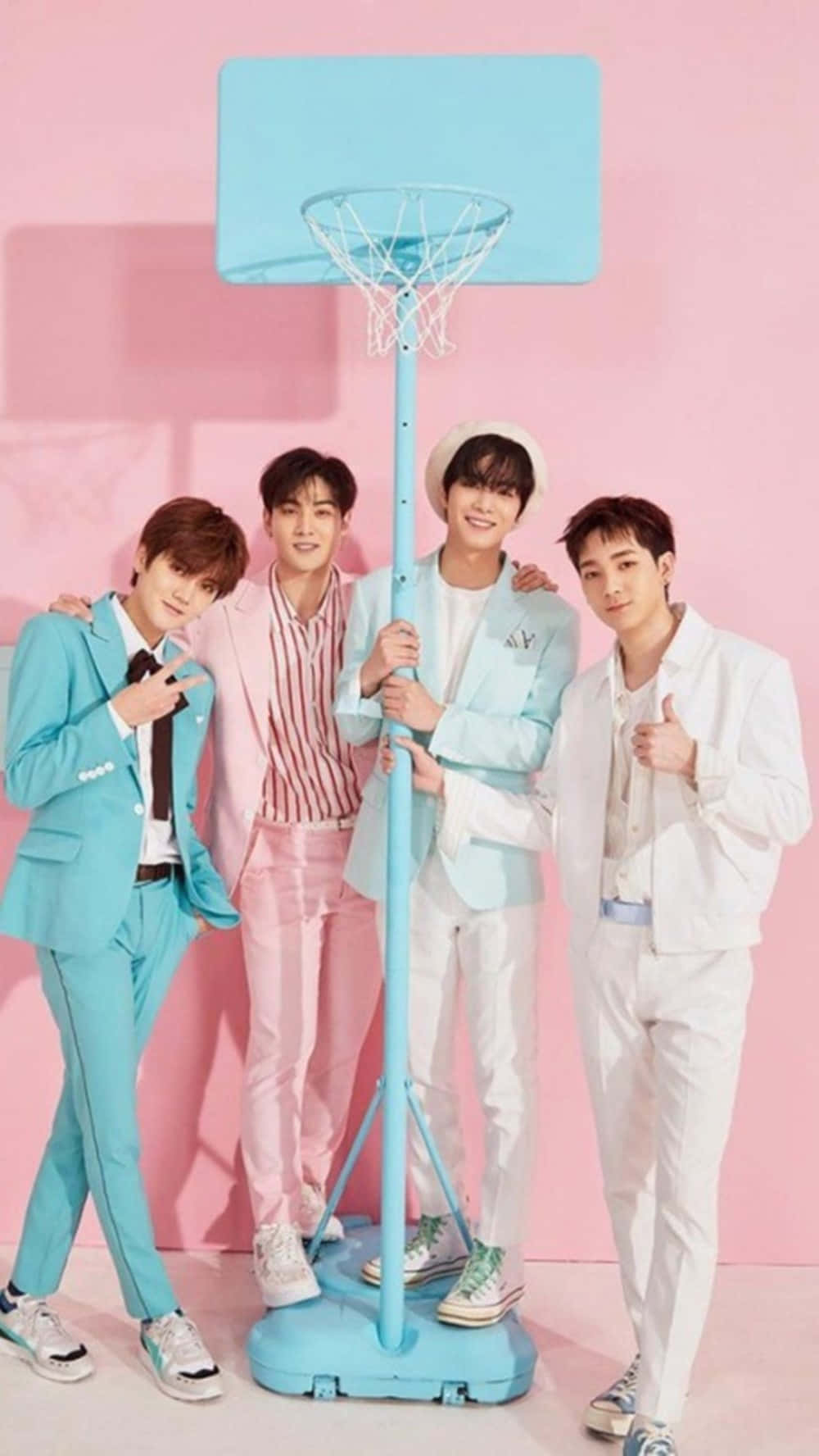 Nuest K-pop 男子團體 Pastel 風寫真 壁紙 — 四位時尚年輕男士身穿粉彩西裝，在充滿活力的粉紅色攝影棚背景前，圍著淺藍色籃球框愉快地擺姿勢。