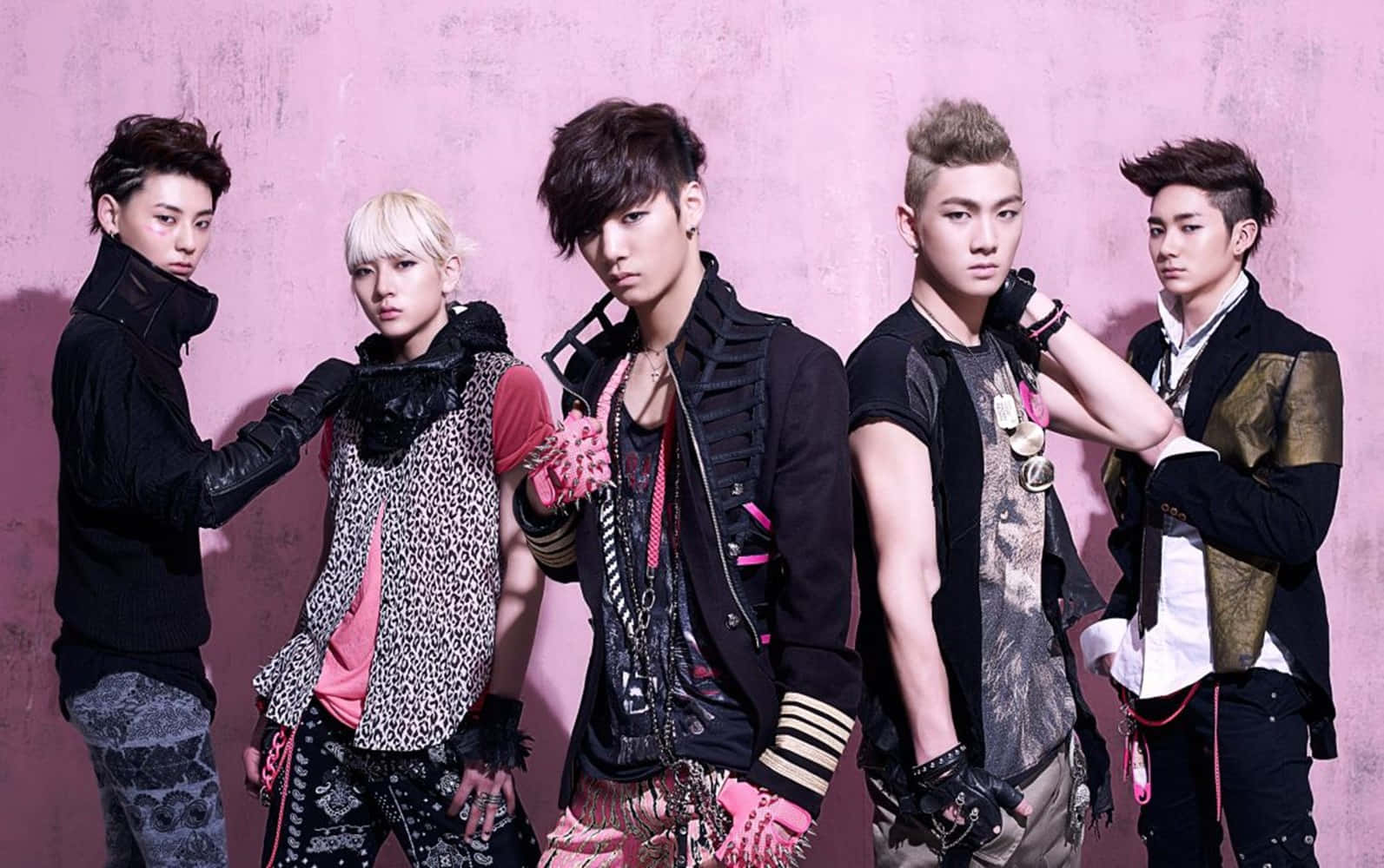 Nuest K-pop 嘻哈風格男子團體 壁紙 — 五位時尚年輕男士穿著前衛服裝，在紋理粉紅牆前自信擺姿勢，表情強烈。