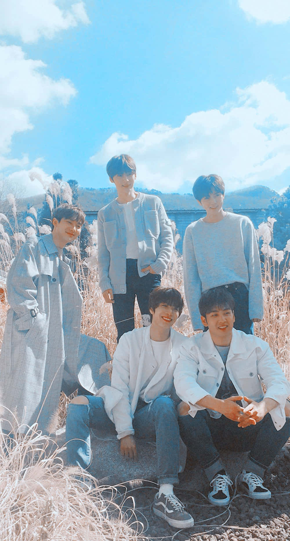 Nuest K-pop 男子團體藍色雲朵編輯照 壁紙 — 五名身穿淺藍色和白色服裝的年輕男子在陽光明媚的高草叢中，在明亮的藍天和蓬鬆的白雲下愉快地擺姿勢。