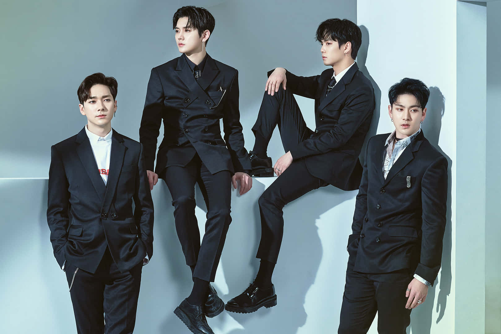 NUEST K-pop男子團體優雅西裝造型 壁紙 — 四位時尚男士身穿利落黑色西裝，在極簡淺藍色工作室牆前自信擺姿勢，帶有戲劇性陰影。