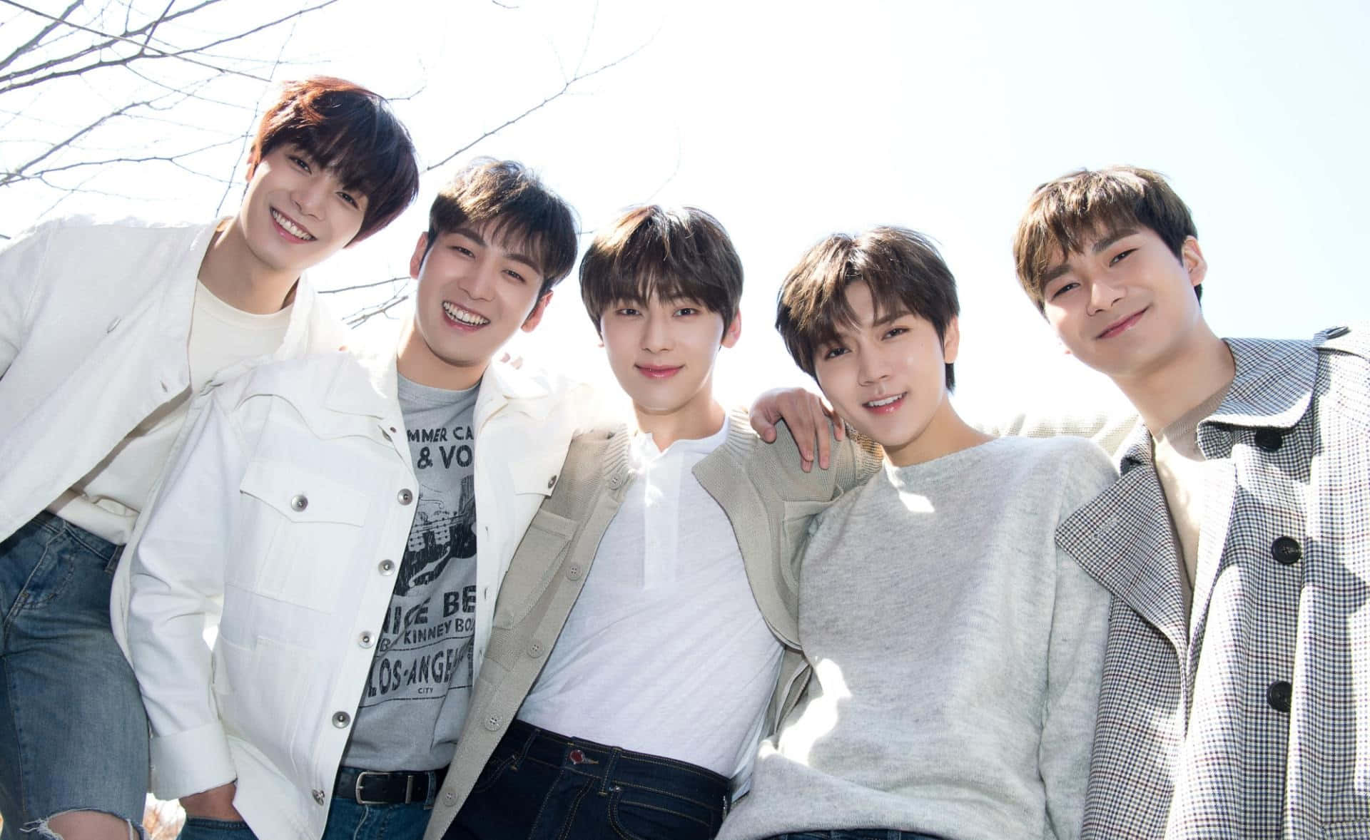 NUEST K-pop男子團體戶外拍攝 壁紙 — 五位微笑的年輕男士穿著休閒春季服裝，在明亮的陽光下緊密地擺姿勢，身後有光禿的樹枝。