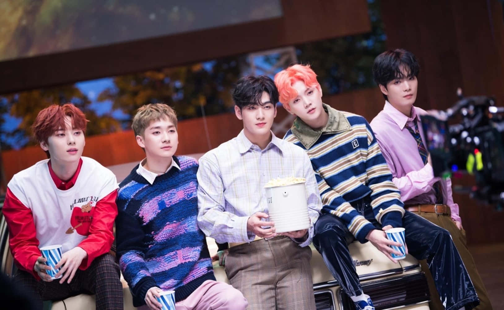 NUEST K-pop男子團體自然拍攝 壁紙 — 五位年輕男子擁有色彩繽紛的頭髮，隨意地坐在場景中，手持爆谷和飲品，穿著時尚休閒服。