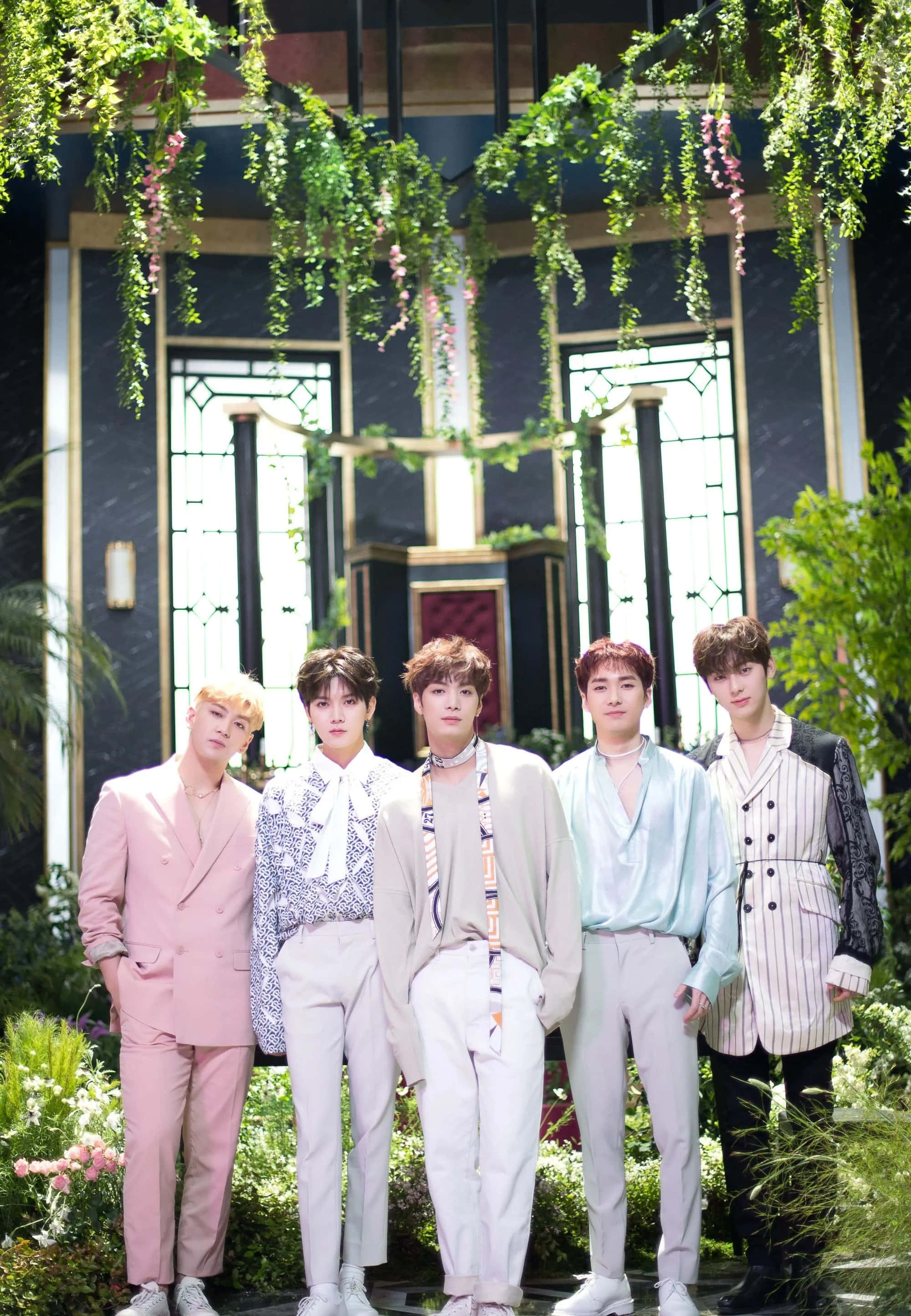 NUEST K-pop男子團體花園主題拍攝 壁紙 — 五位穿著粉彩西裝的時尚年輕男子在陽光普照的花園庭院中，在茂密的懸掛綠植下自信地擺姿勢。