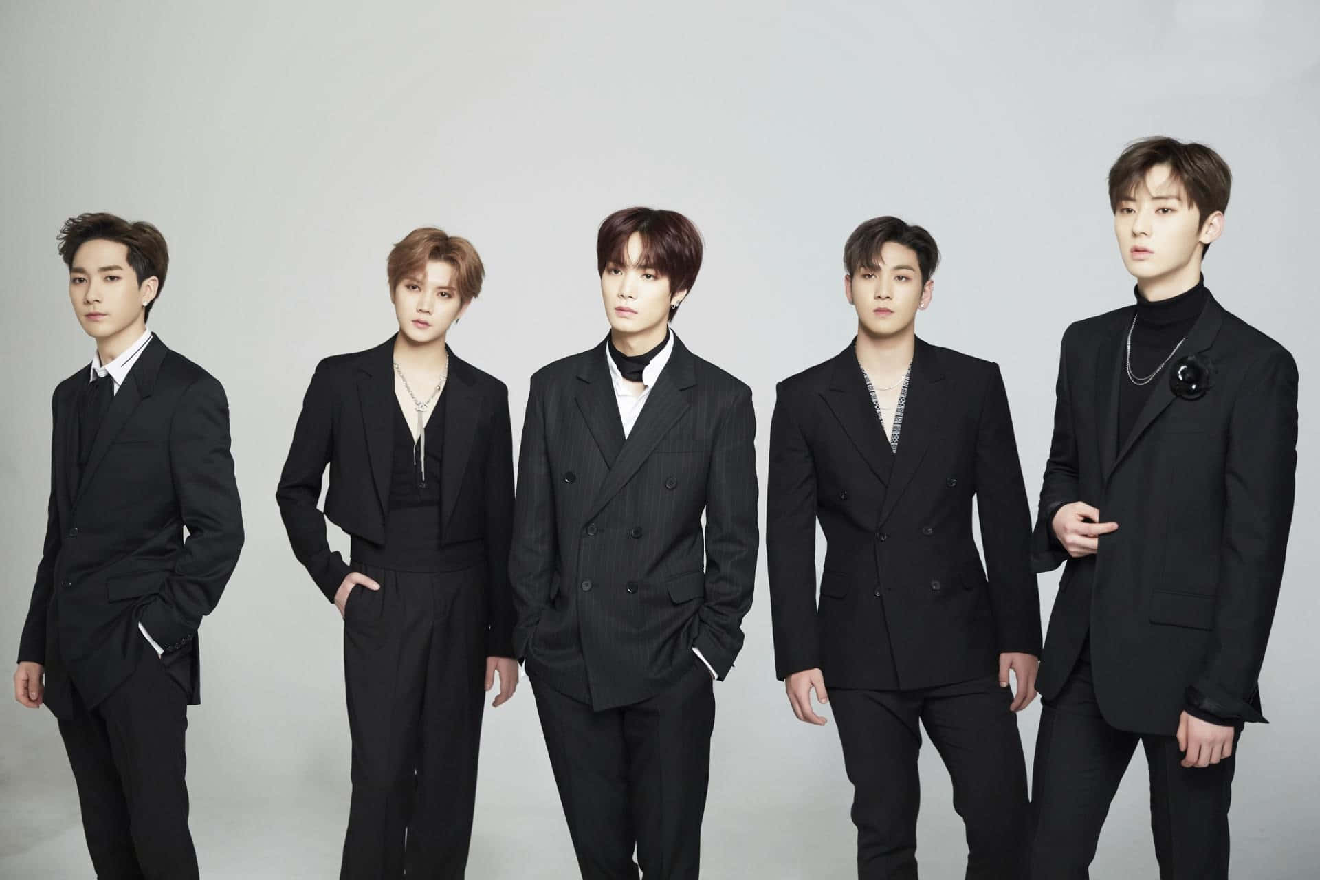NUEST KPOP PLEDIS 廣告拍攝 壁紙 — 五位時尚年輕男士身穿利落黑色西裝，在純灰色攝影棚背景前自信地擺姿勢。
