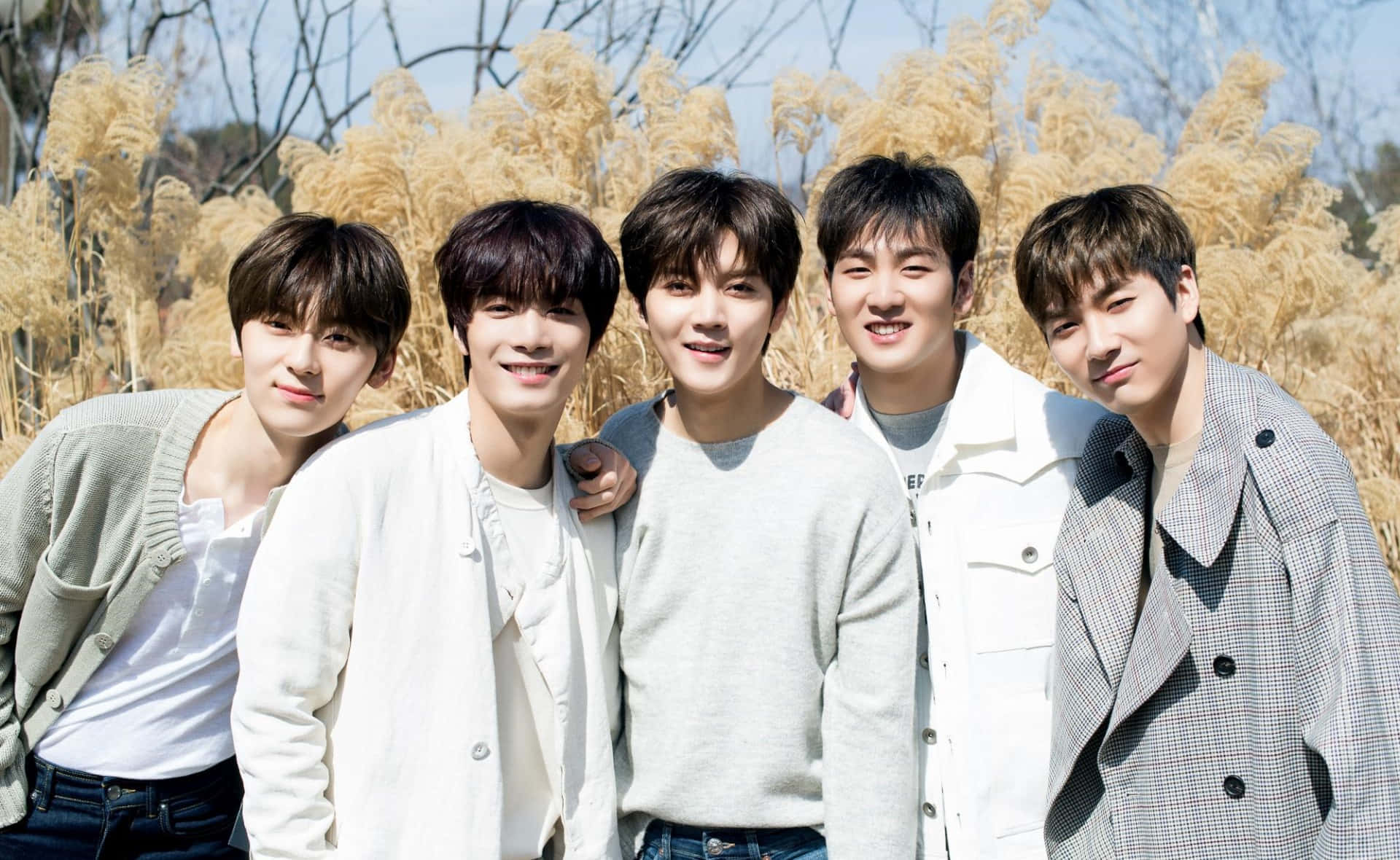 NUEST KPOP 男子團體成員 壁紙 — 五位微笑的年輕男性穿著休閒外套，在清澈藍天下以高大的金色蒲葦為背景緊密站在一起。