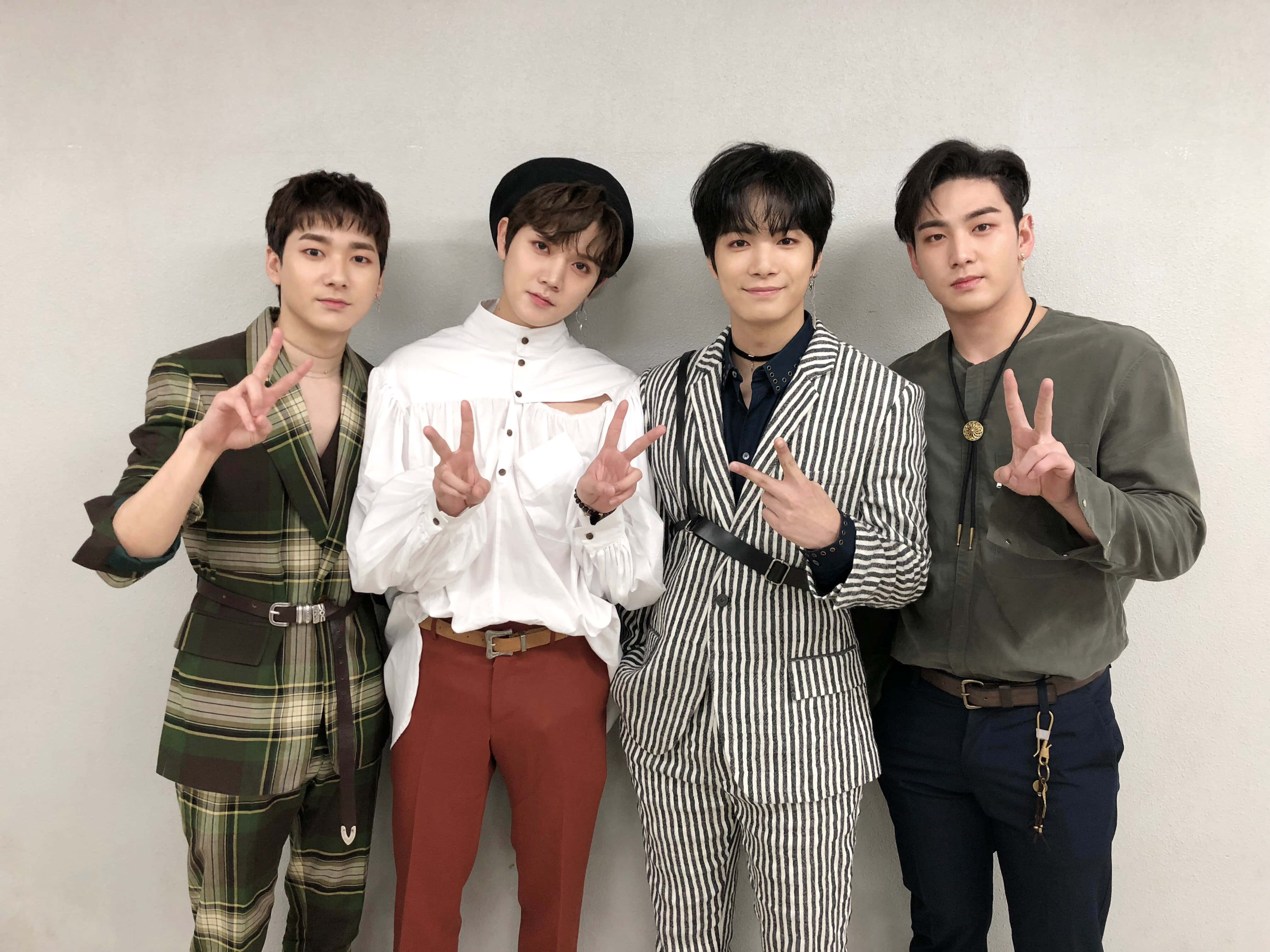 NUEST KPOP 男子團體演出前照 壁紙 — 四位時尚年輕男士穿著潮流服裝，自信地靠著素牆擺姿勢，每人雙手做出和平手勢。
