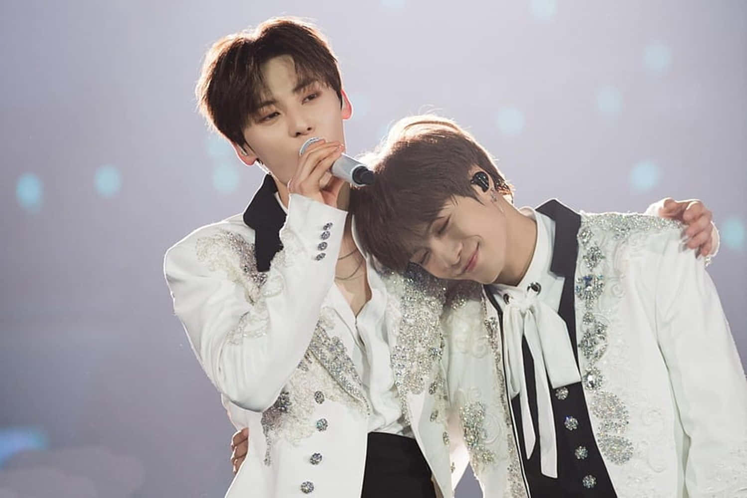 Nuest 男子團體演唱會現場照片 壁紙 — 兩位身穿白色閃亮外套的男表演者在柔和藍光下於舞台上分享親密時刻。