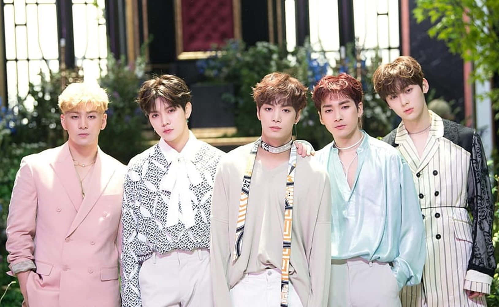NUEST古典風造型K-pop男子團體 壁紙 — 五位穿著粉彩和圖案服裝的時尚年輕男子在茂密花園背景下自信地戶外擺姿勢。