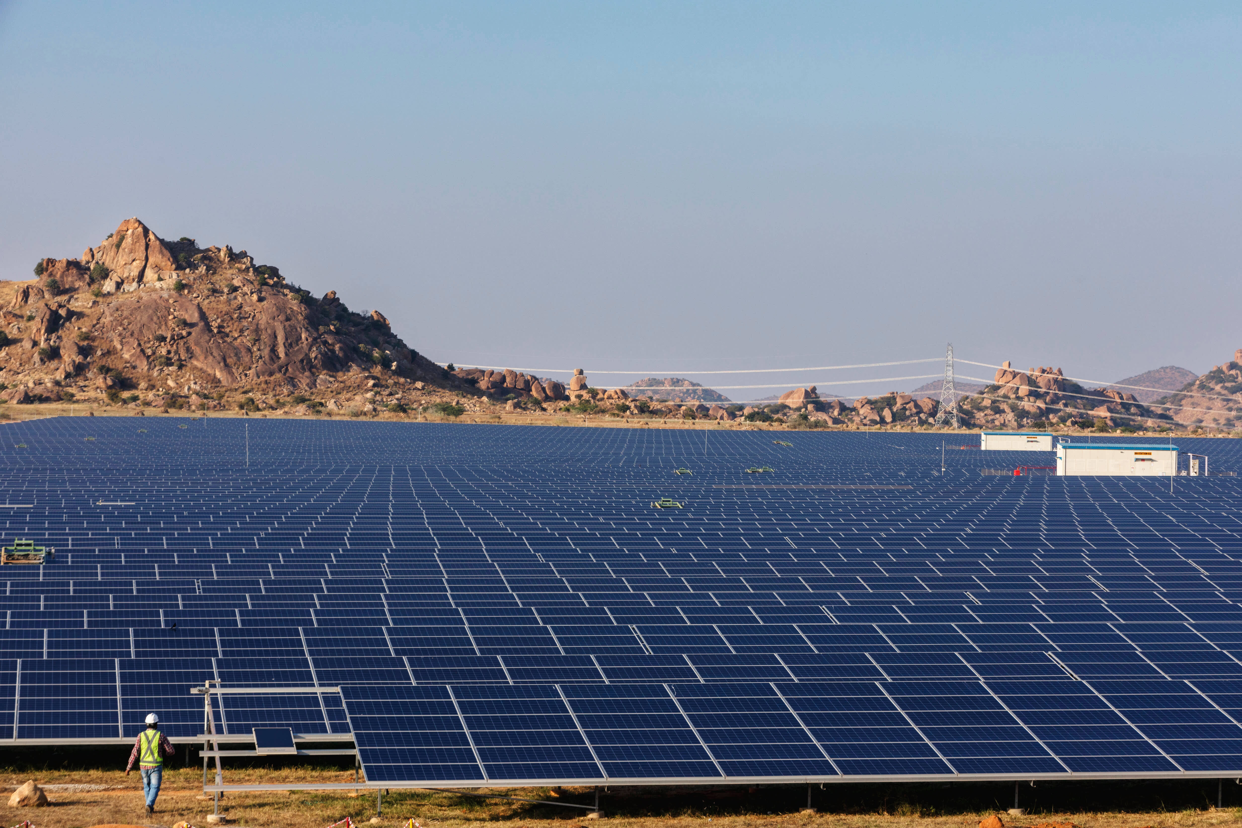 NV Energy：創造更光明的未來 壁紙 — 一名孤獨的工人在晴朗的天空下，在廣闊的藍色太陽能板之間行走，背景是岩石丘陵。