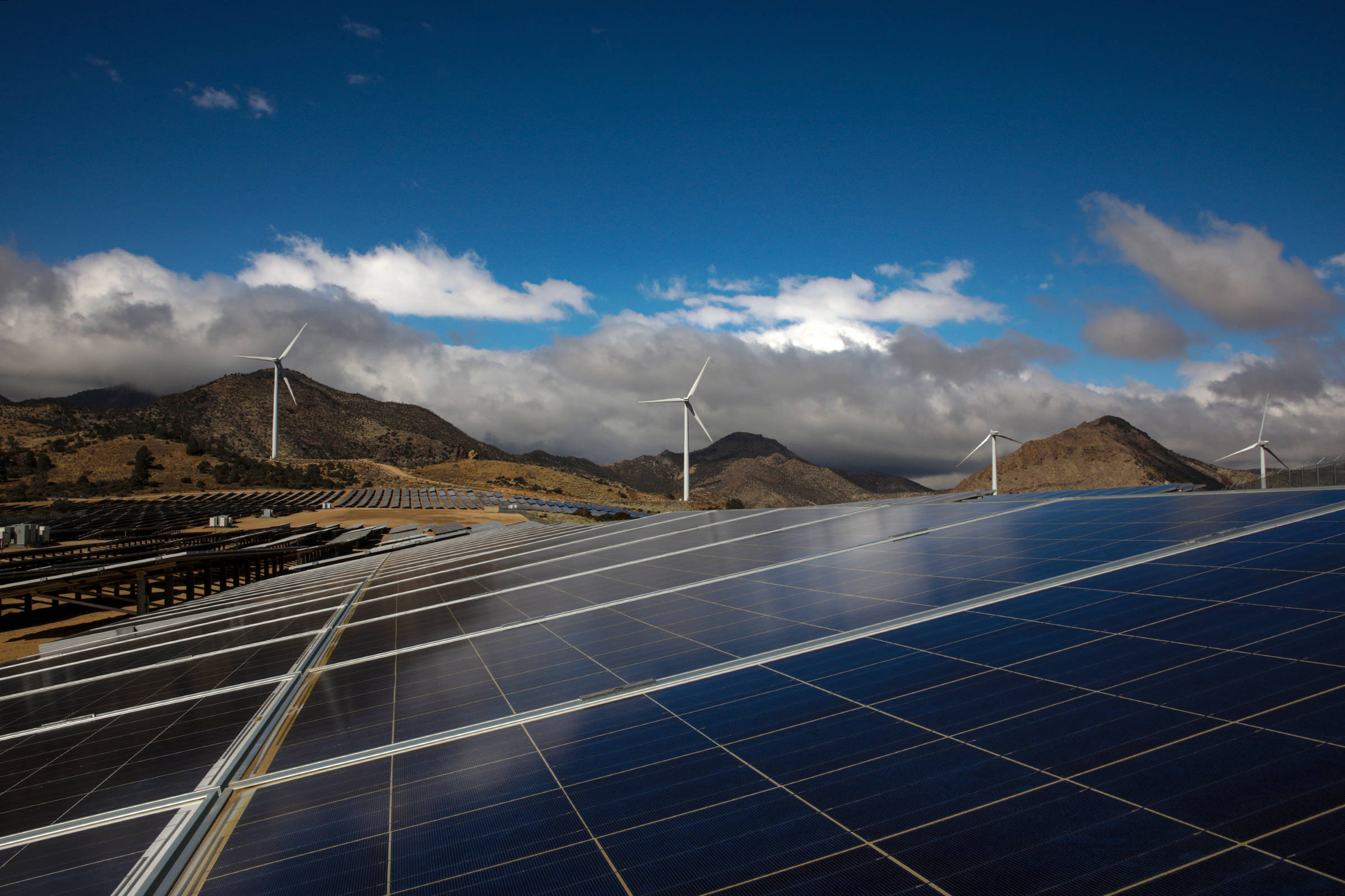 NV Energy 電力來源 壁紙 — 一排排太陽能板在陽光明媚的沙漠景觀中延伸，風力渦輪機在連綿起伏的山丘後方升起，天空多雲，藍天白雲。
