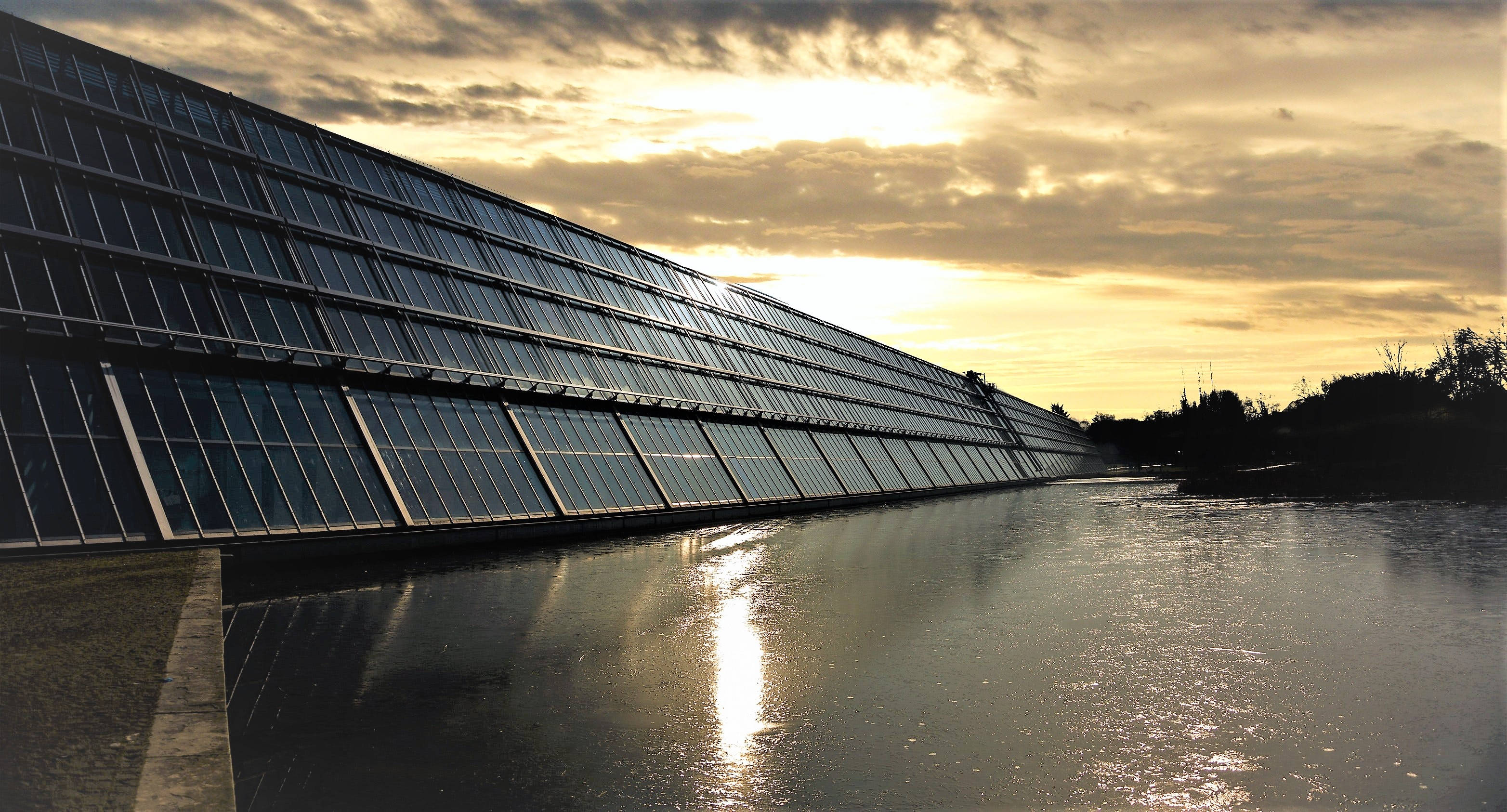 NV Energy水庫壁掛太陽能板 壁紙 — 一座現代玻璃建築在平靜的水面上反射出金色夕陽的光芒，天空中有戲劇性的雲彩。