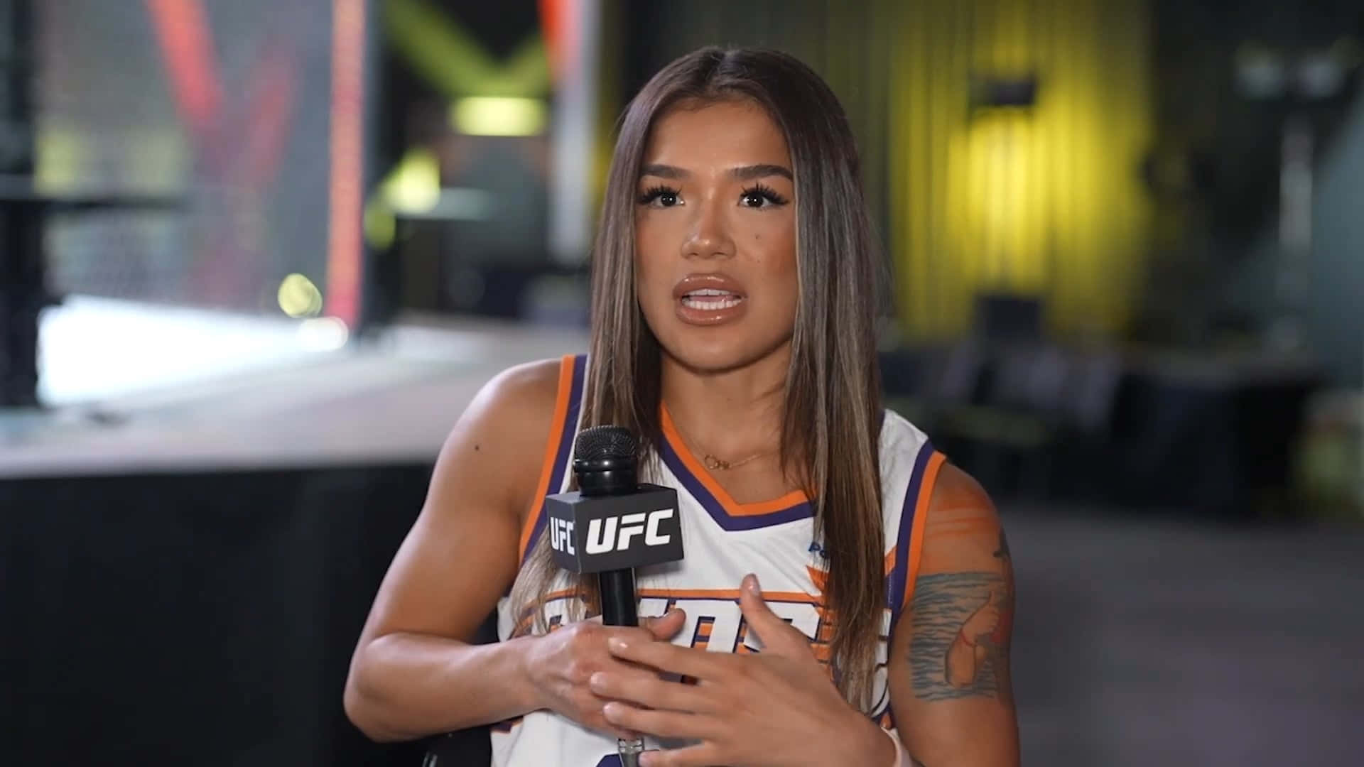  壁紙 — 一位身穿白色、橙色和藍色球衣的女性UFC拳手，神情專注地對著麥克風說話。